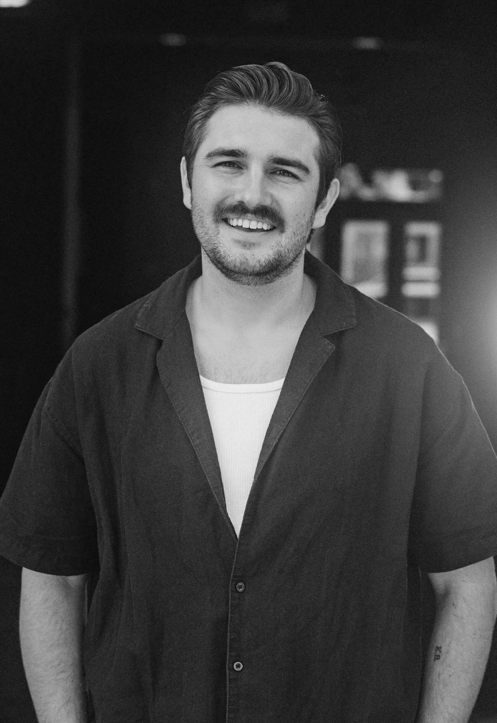 A smiling young man with dark hair and a beard, wearing a black shirt over a white t-shirt, standing outdoors during sunset.