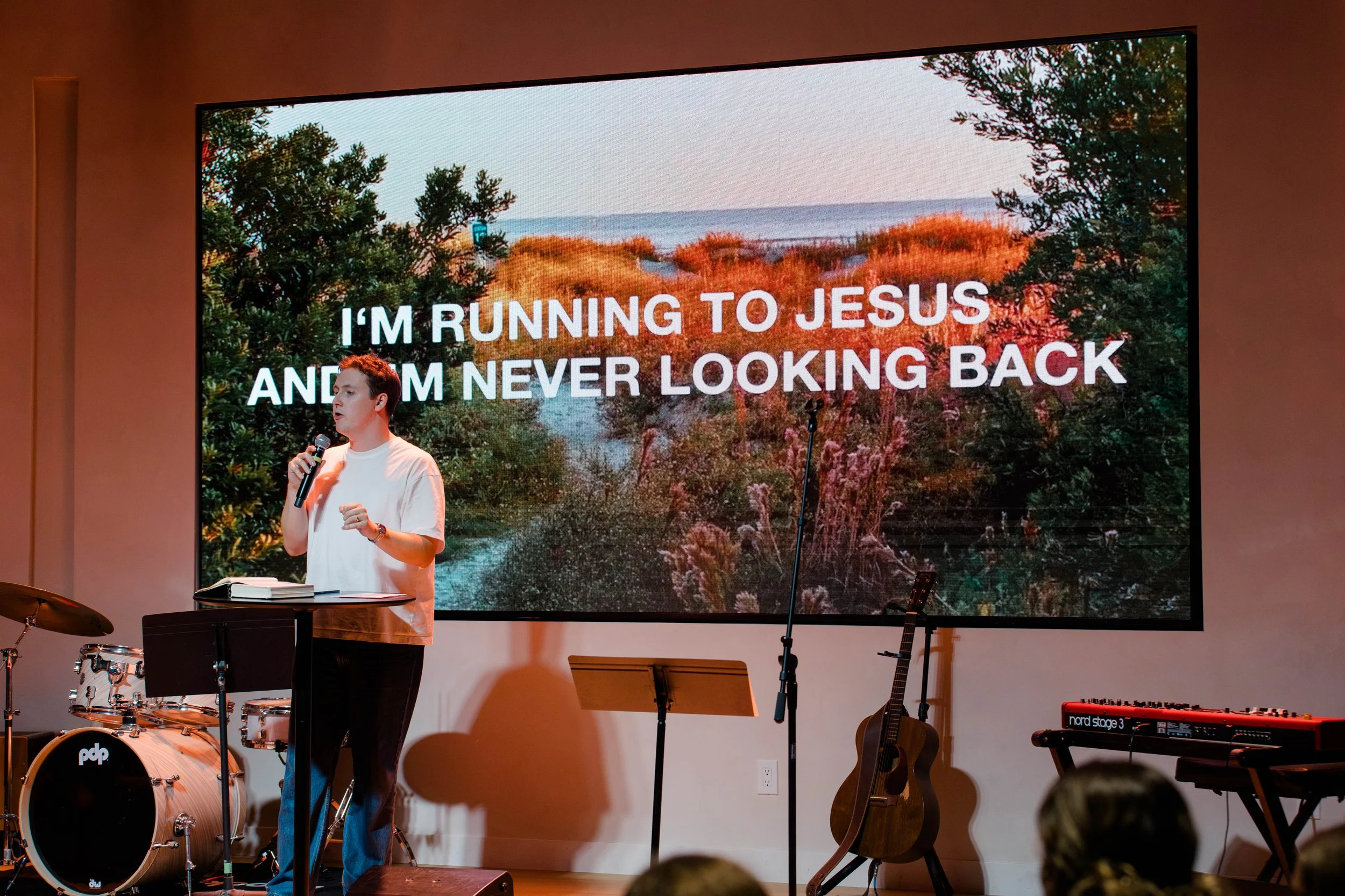 A young man holding a microphone standing in front of a large screen displaying nature scenery and the text "I'M RUNNING TO JESUS AND I'M NEVER LOOKING BACK" in a church or worship setting. Musical instruments are visible on stage.
