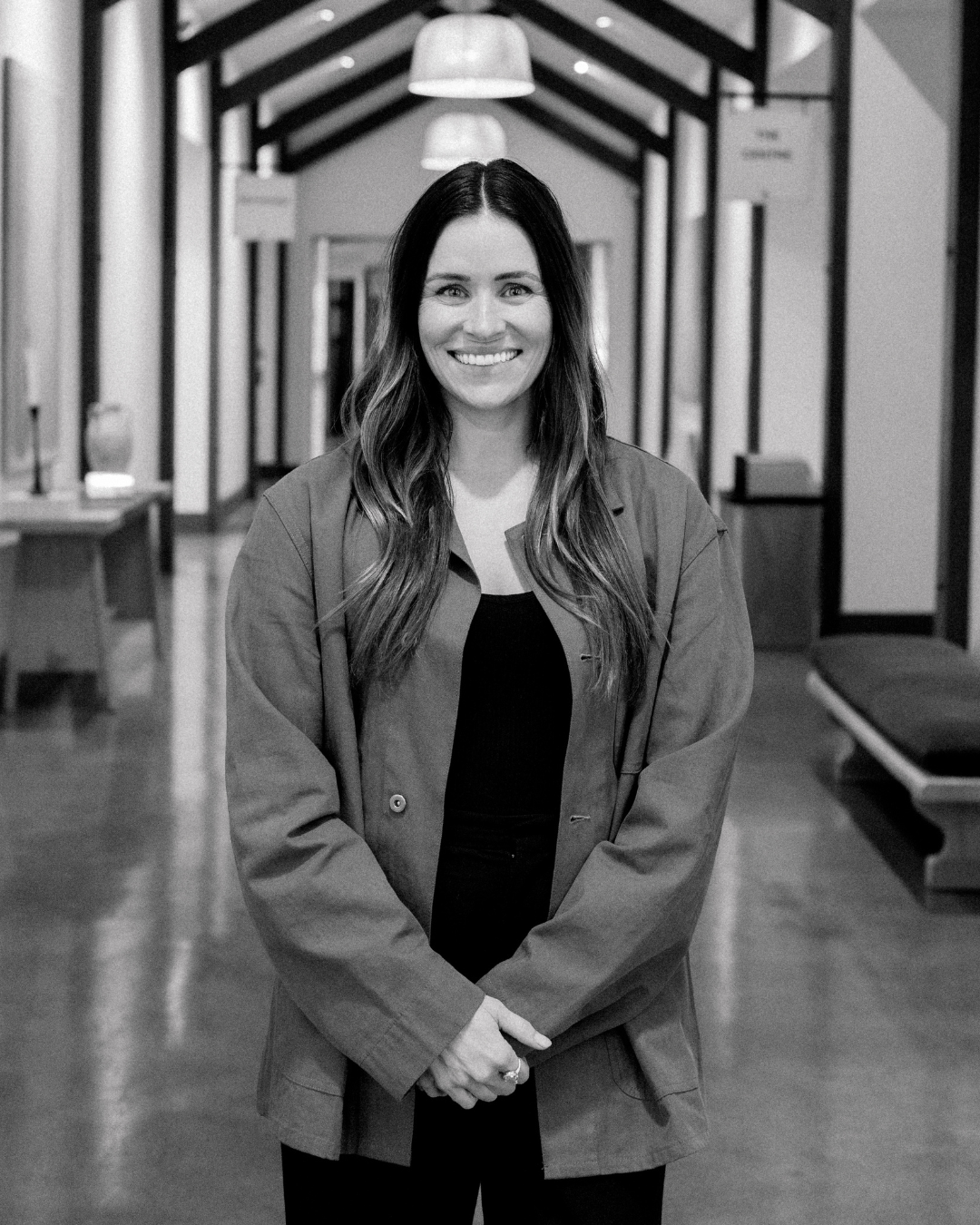 A woman with long, curly brown hair smiling outside during sunset, wearing a denim jacket over a white top.