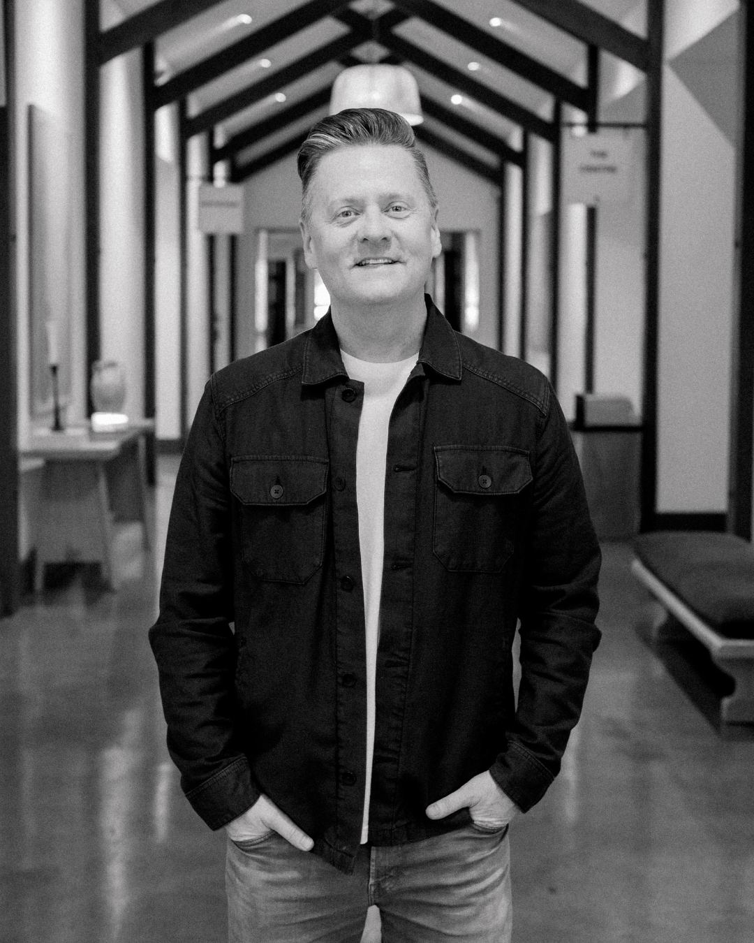 A man with light skin, short light brown hair, and a slight smile stands outdoors in front of a dark background with sunlight on the right side of the image. He is wearing a denim jacket over a beige t-shirt.