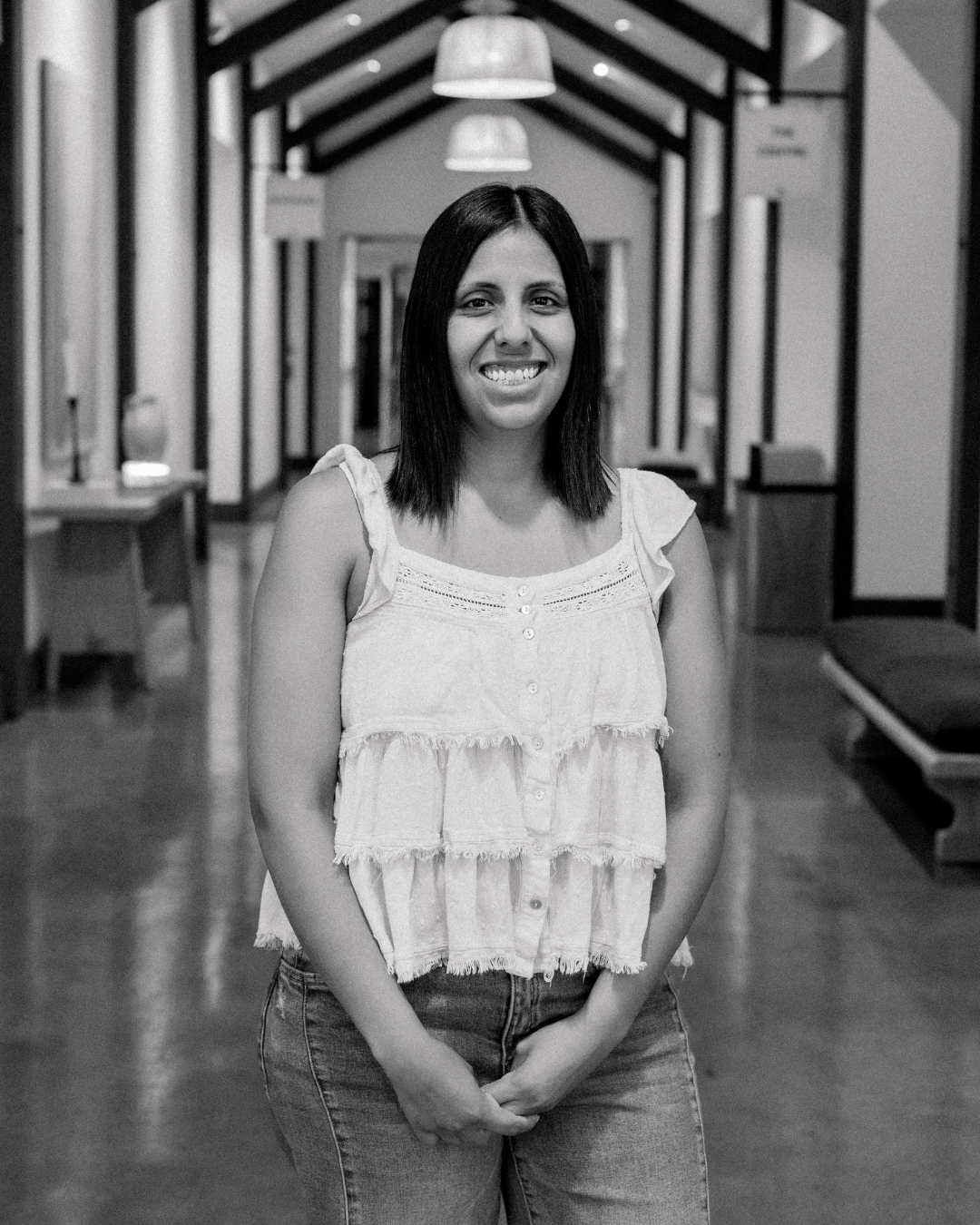 A woman with shoulder-length dark hair smiling and wearing a light denim shirt over a mauve t-shirt, standing indoors with windows in the background.