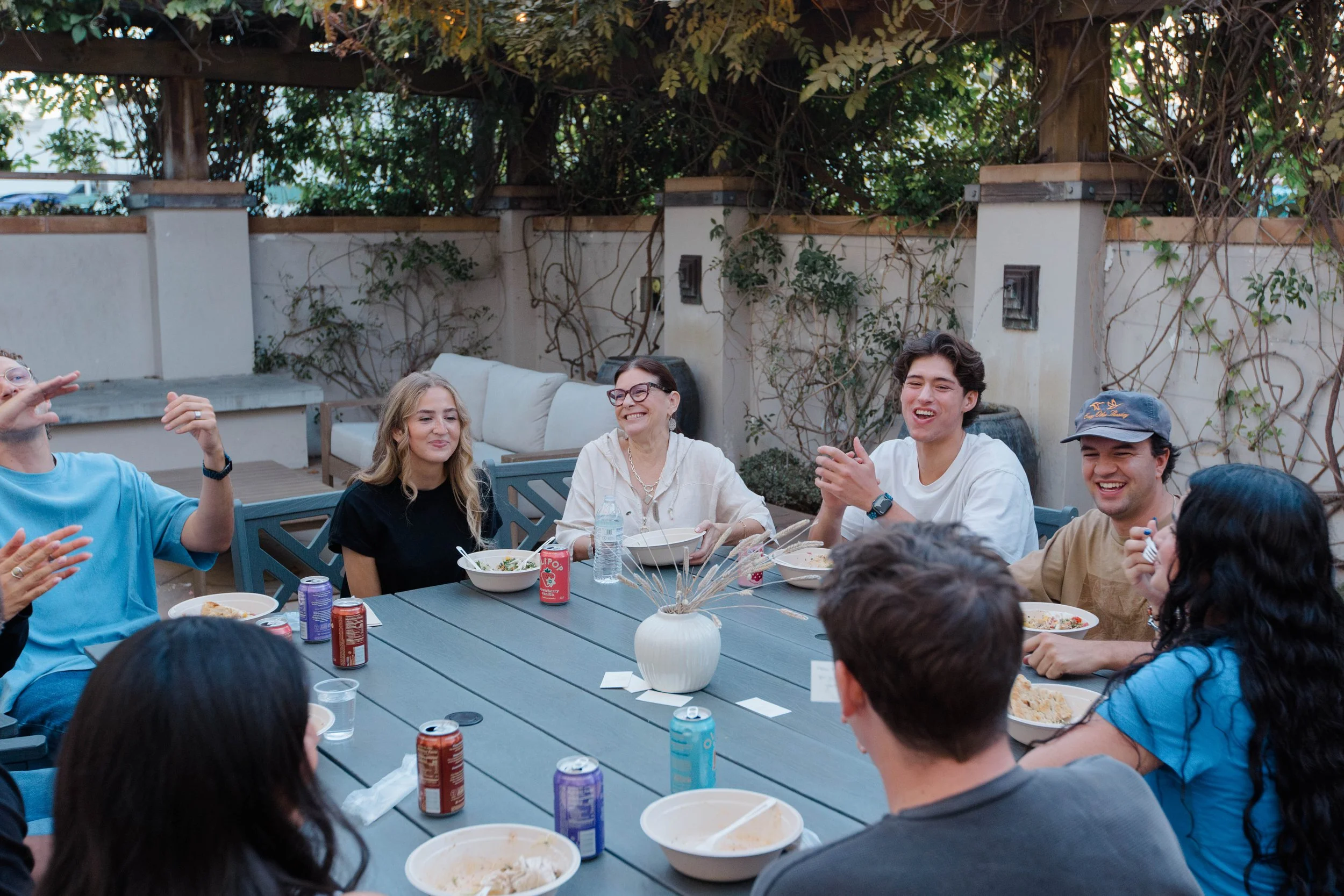 People sitting at a long table eating food, with a plant centerpiece, in an indoor setting with wood-paneled walls.