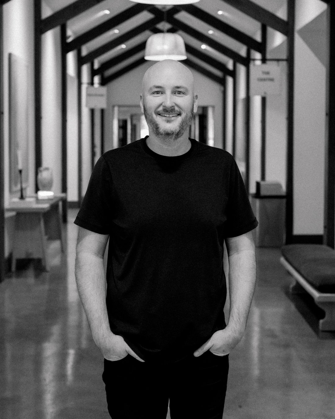 Smiling man with a shaved head and beard, wearing a gray button-up shirt, standing outdoors during sunset.