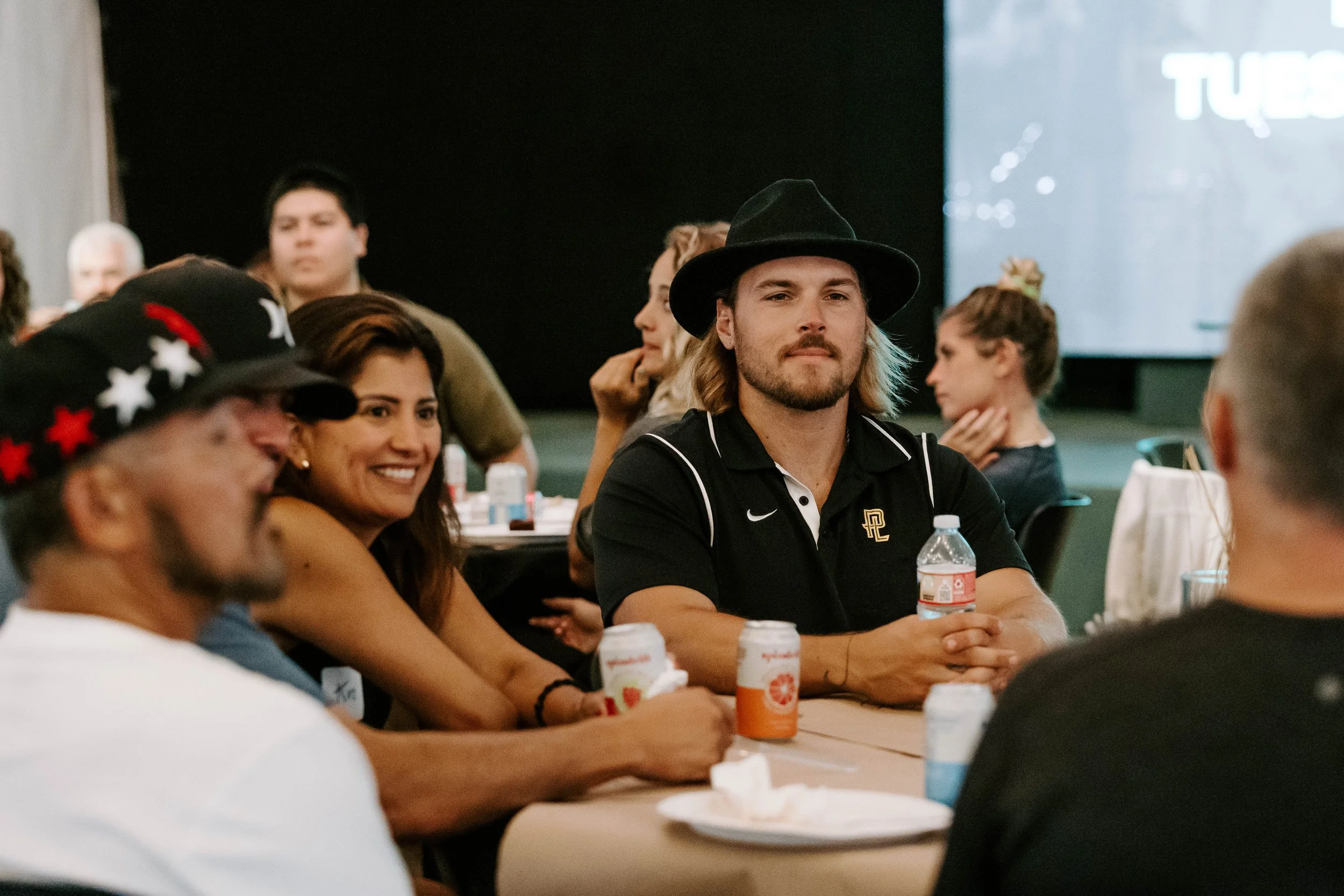 A group of people sitting at a table with drinks during an indoor event, including a man wearing a black hat and a black polo shirt, a smiling woman, and others in the background.