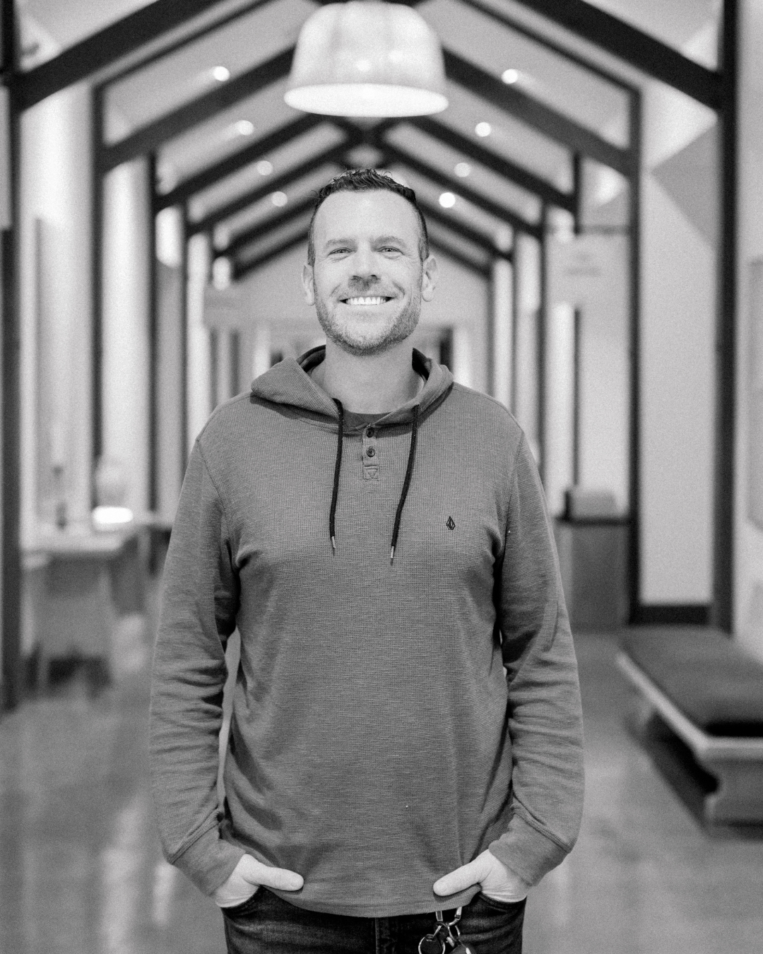 A smiling man with short brown hair and a beard, wearing a white T-shirt with the word 'OBEYN' and a logo, standing outdoors during sunset.