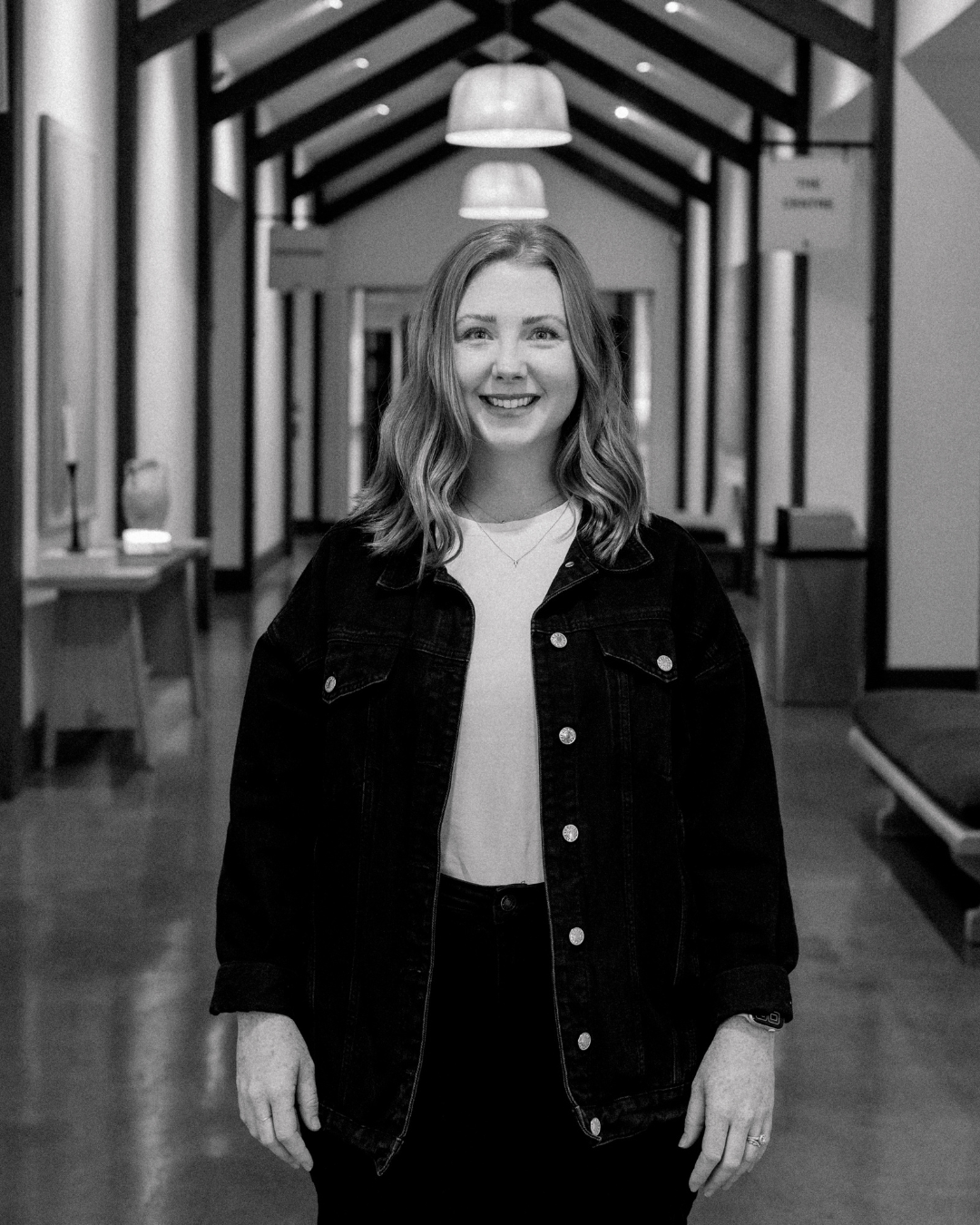 A young woman with long red hair smiling, wearing a white shirt and a brown checkered blazer, standing indoors with warm lighting.