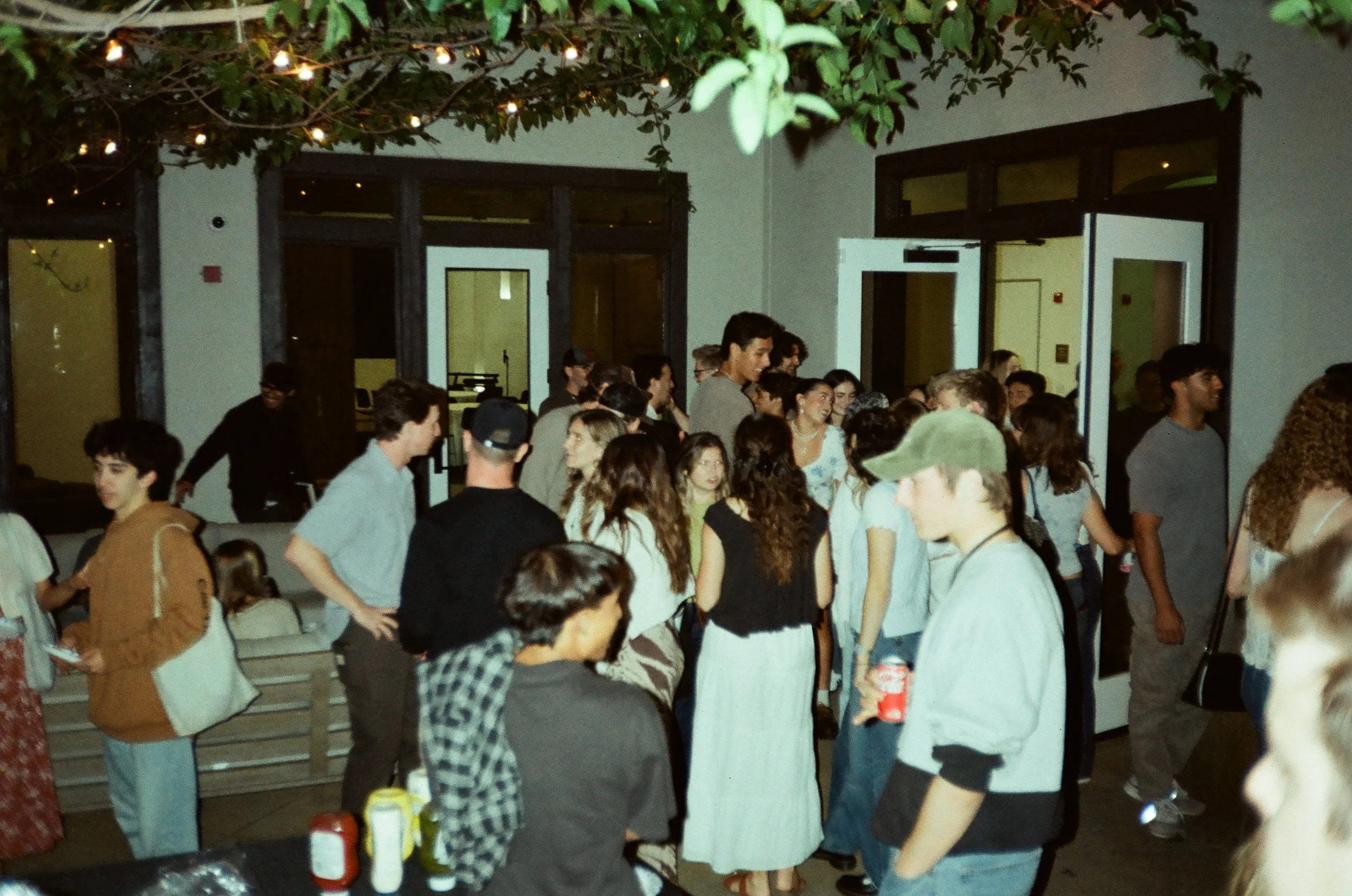 A large group of people gathered indoors and outside, socializing and enjoying an event during the evening. The scene includes visible drinks, condiments, and string lights hanging above, creating a lively atmosphere.