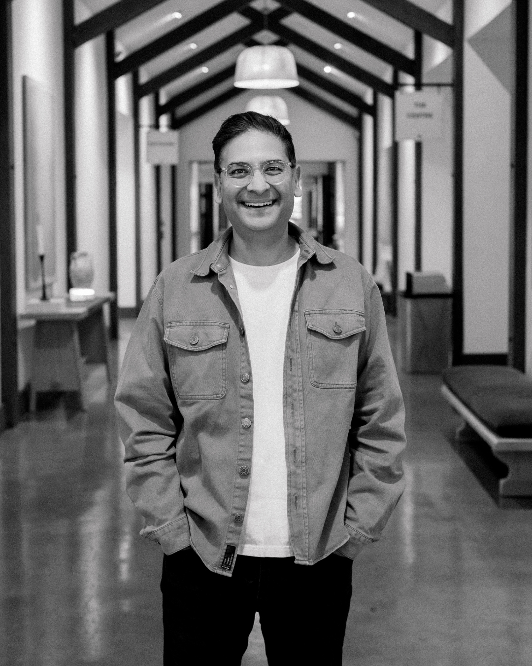 A man standing with arms crossed, wearing glasses and a black jacket inside a well-lit room with wooden furniture and large windows.