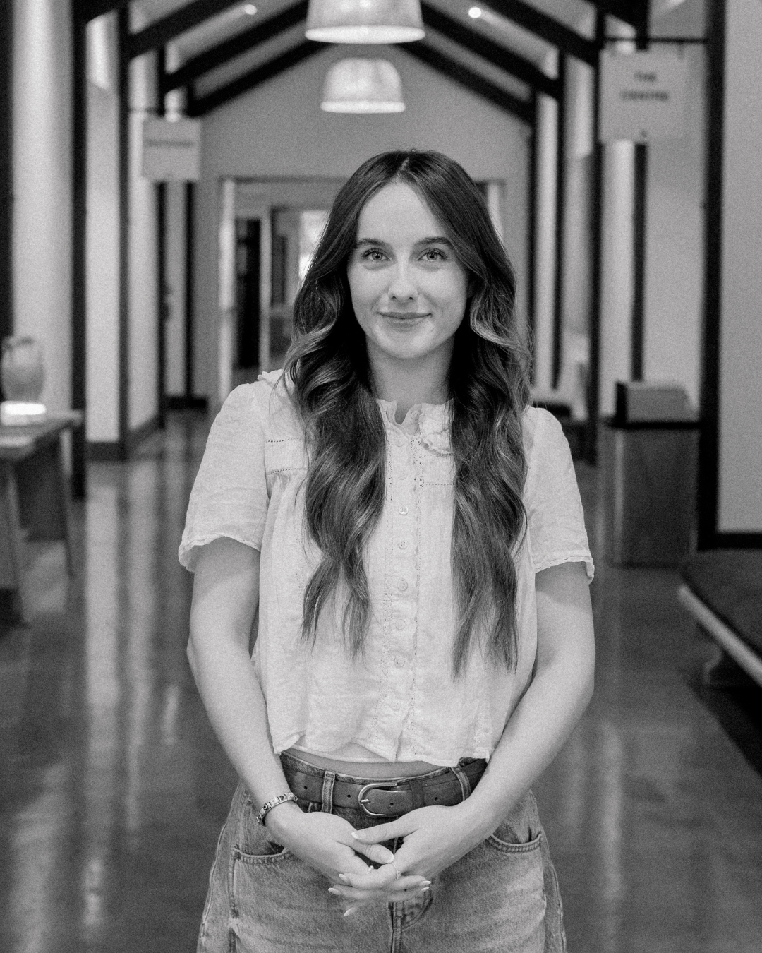 A woman with long brown hair wearing a white blouse, smiling indoors.