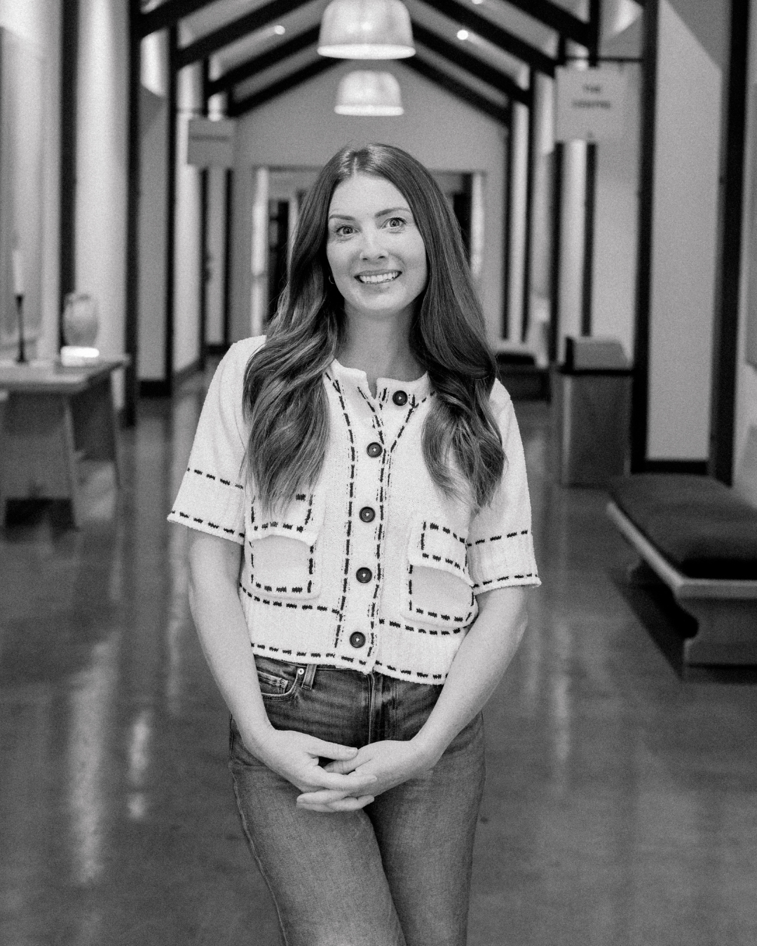 A woman with long red hair smiling outdoors, wearing a white button-up shirt over a light gray top.