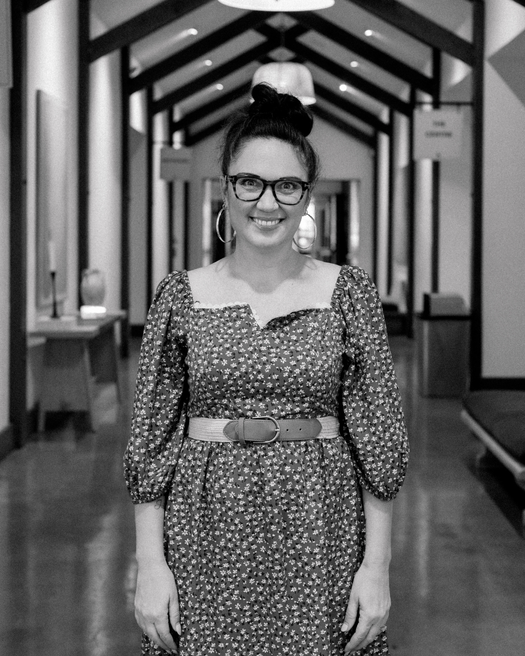 A smiling woman with long wavy brown hair and glasses, wearing a black dress with floral embroidery, standing outdoors during sunset.