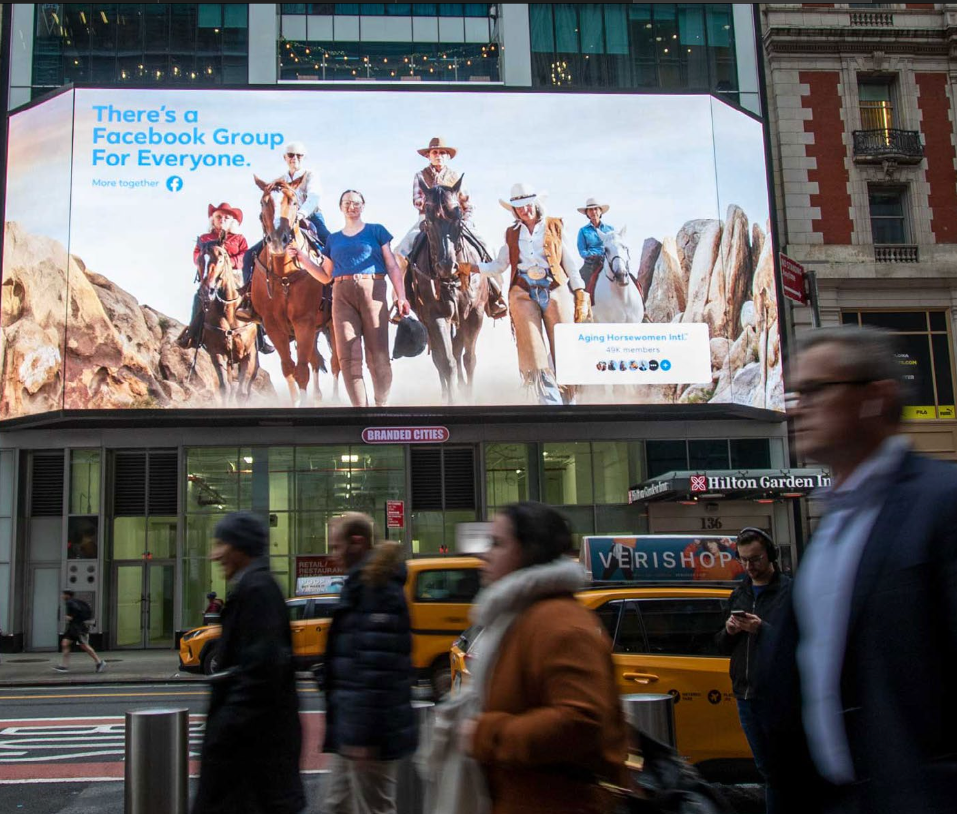 Large electronic billboard displaying an advertisement for Facebook's group 'Aging Horsewomen Intl.' featuring women on horseback in a desert landscape, with the text 'There's a Facebook Group for Everyone.' Below the billboard, a busy city street scene with pedestrians and yellow taxis.