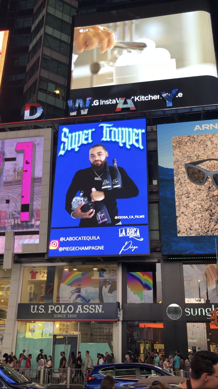 Billboard advertisements in Times Square, featuring a large screen showing a woman cooking, a digital ad for Super Truper with a man holding bottles, U.S. Polo Assn. store, and other billboards with shops and pedestrians below.