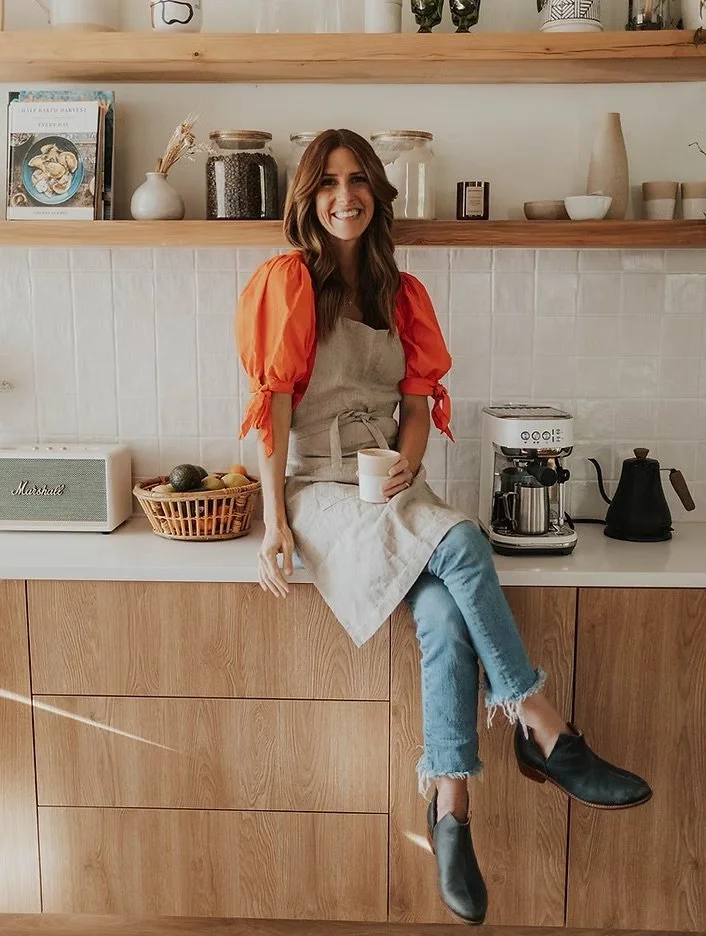 Woman sitting on kitchen counter, wearing a red-sleeved apron, holding a mug, with a coffee maker and fruit basket nearby.