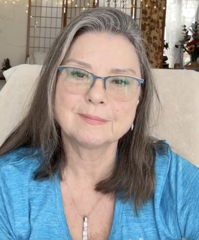 Woman with glasses and long hair wearing a blue shirt, indoors with decorative background.