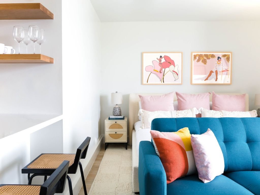Modern living room with a blue sofa, decorative pillows in various colors, floating shelves with wine glasses, framed art on the wall, and a small side table with a lamp.