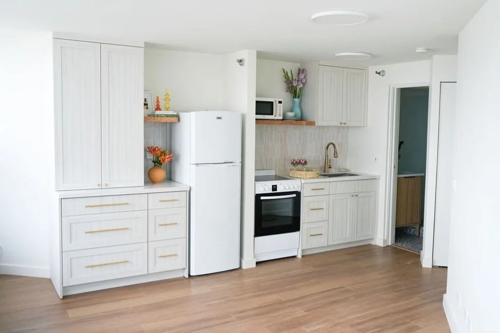 Compact modern kitchen with white cabinets, fridge, stove, oven, and sink; wooden floors and minimal decor.