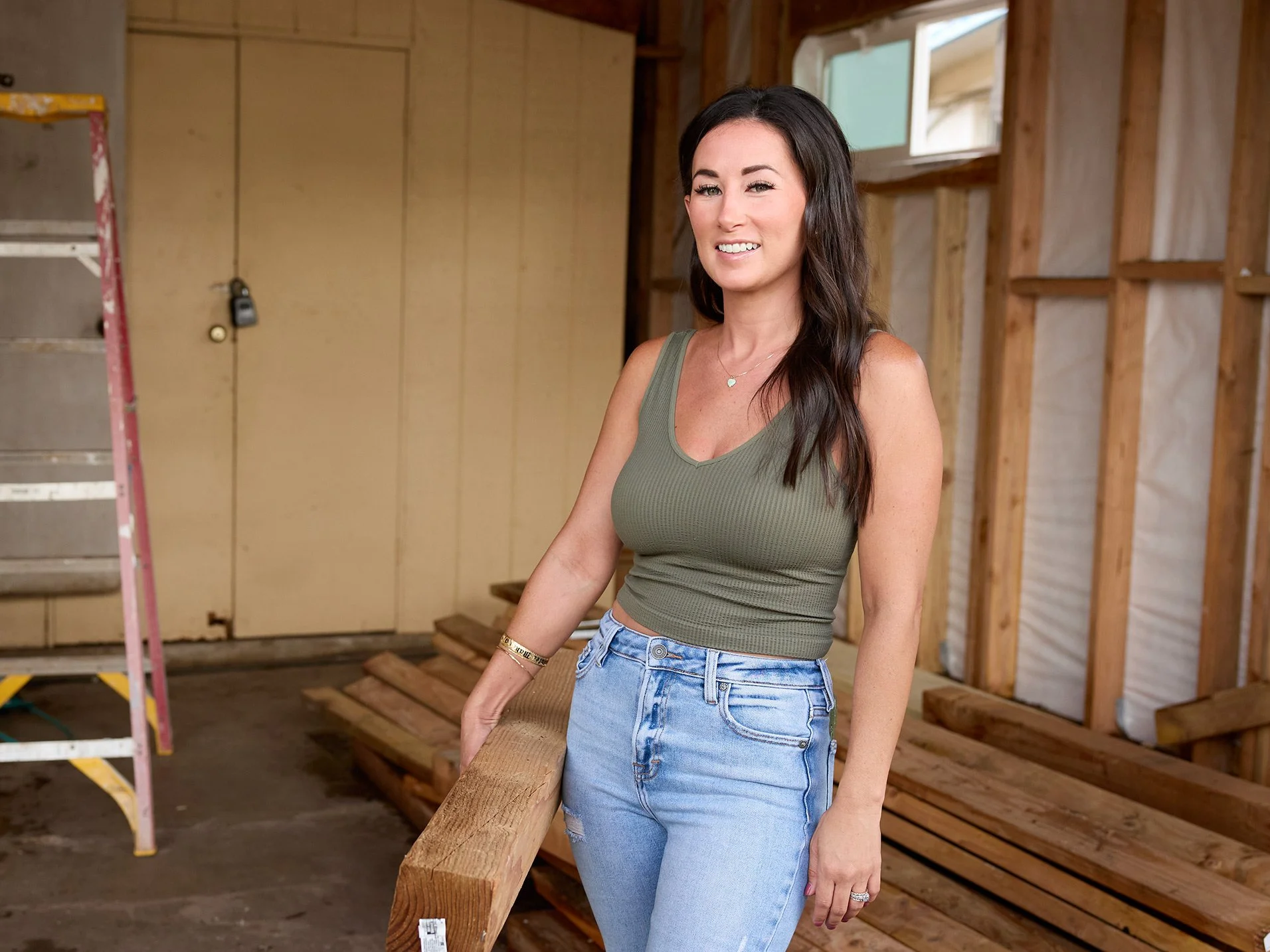 A woman in a green tank top and jeans stands in a construction area with wooden beams and a ladder in the background.