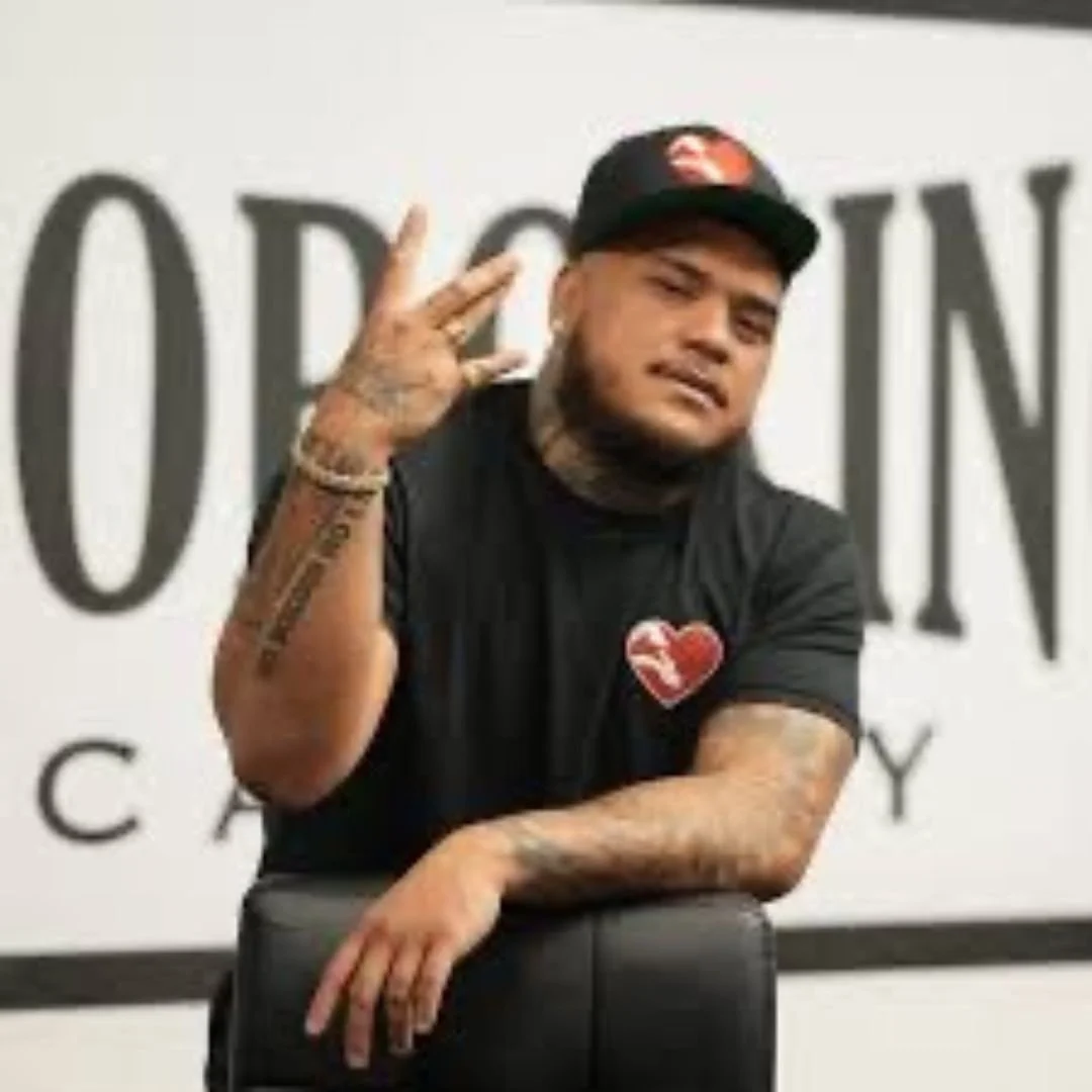 Person wearing a black cap and t-shirt with heart symbol, showing a hand gesture in front of a blurred background with text.