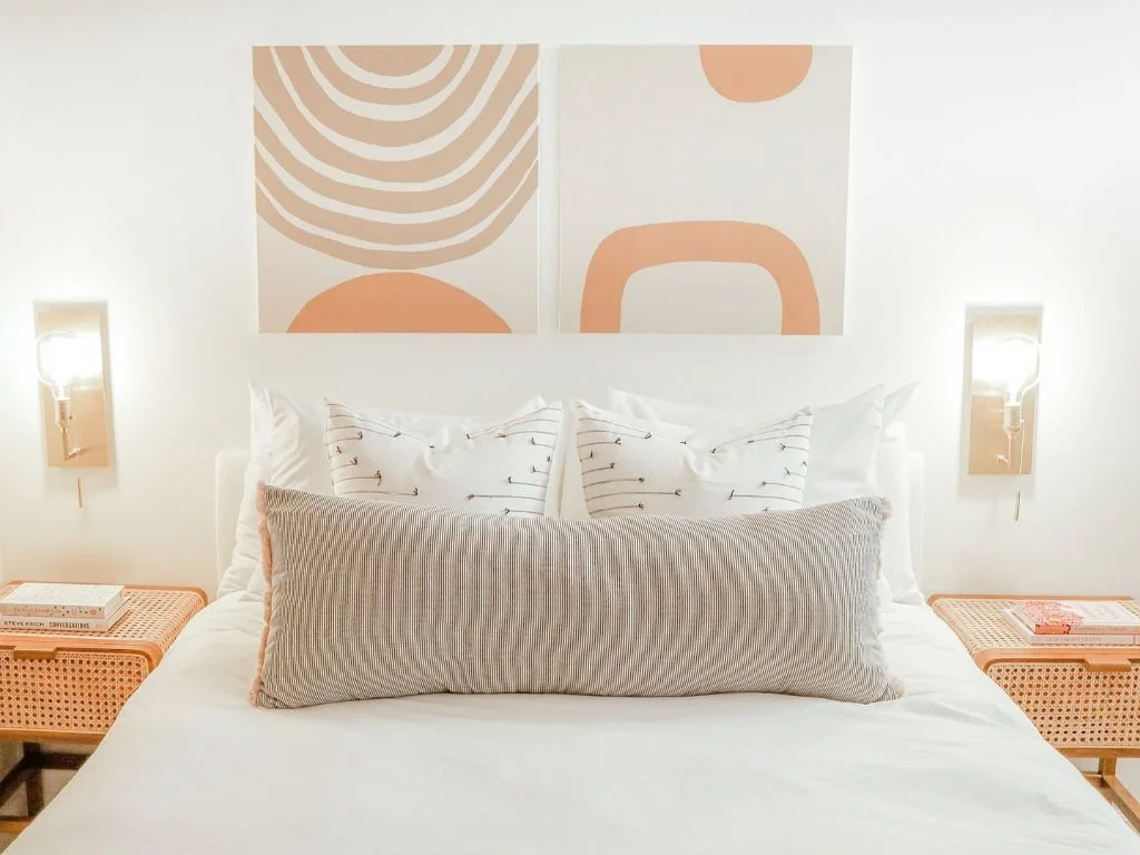 Modern bedroom interior with a white bed, decorative pillows, minimalist abstract art, and wall-mounted lights. Two wicker nightstands with books flank the bed.