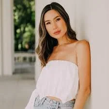 Woman in white top and denim shorts leaning against a wall outdoors.