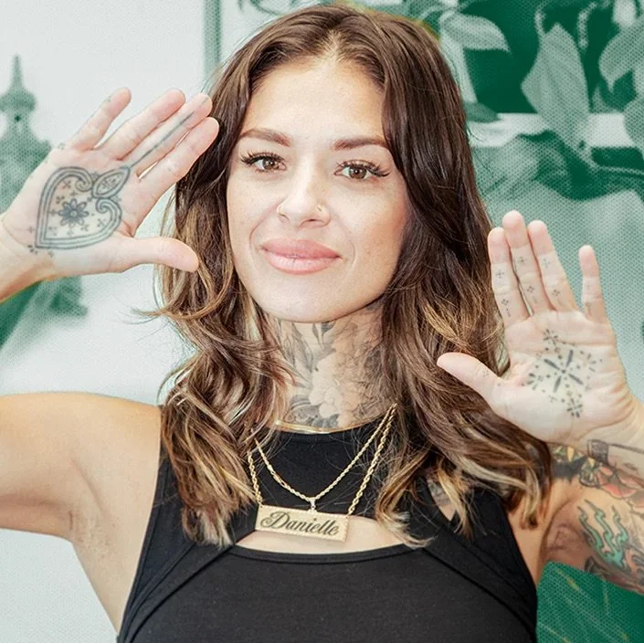Woman with tattoos raising both hands, showing palm tattoos, wearing a necklace and a black top, smiling.