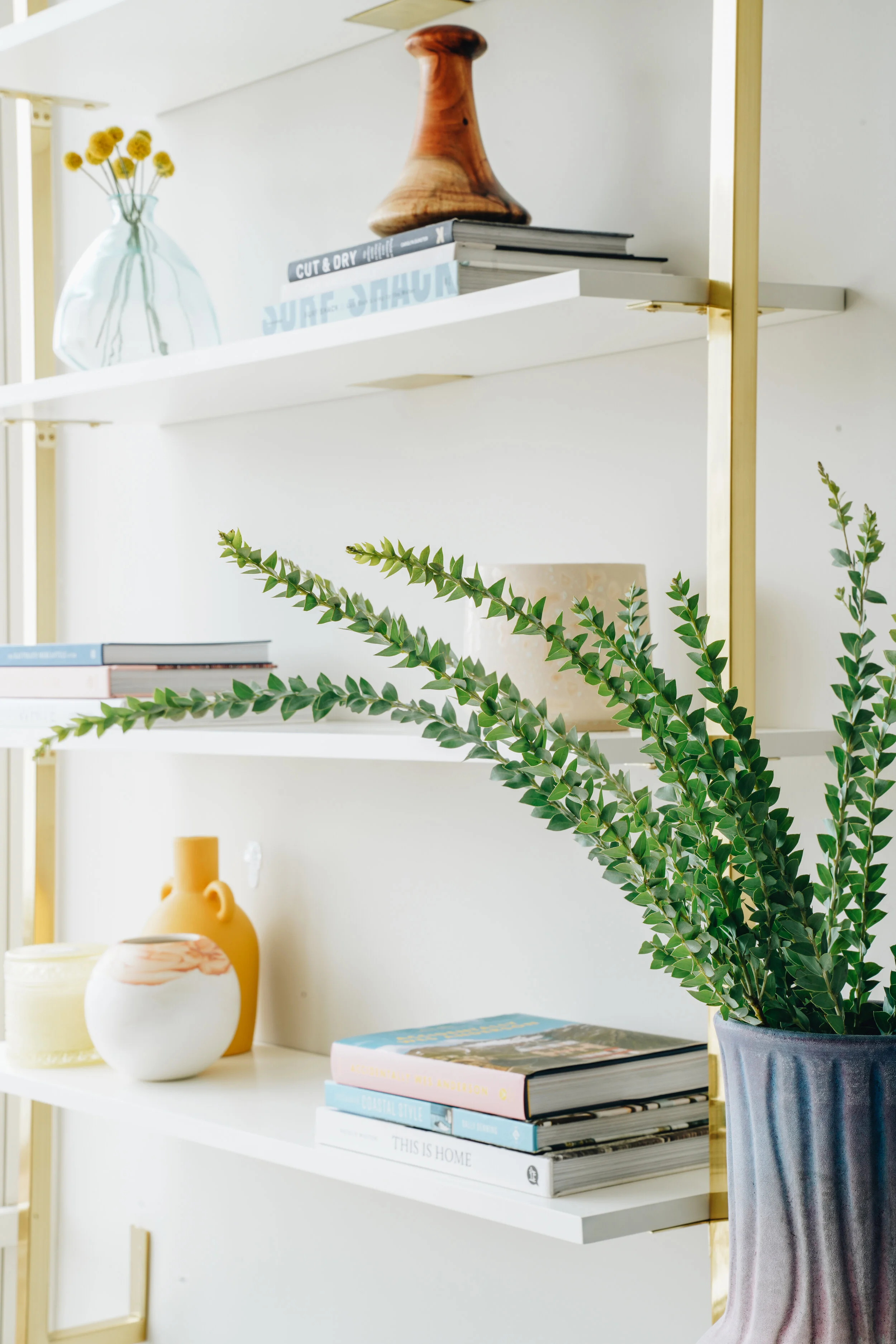 Modern bookshelf with decorative items, including potted plant, books, vases, and wooden sculpture on white shelves with gold supports.