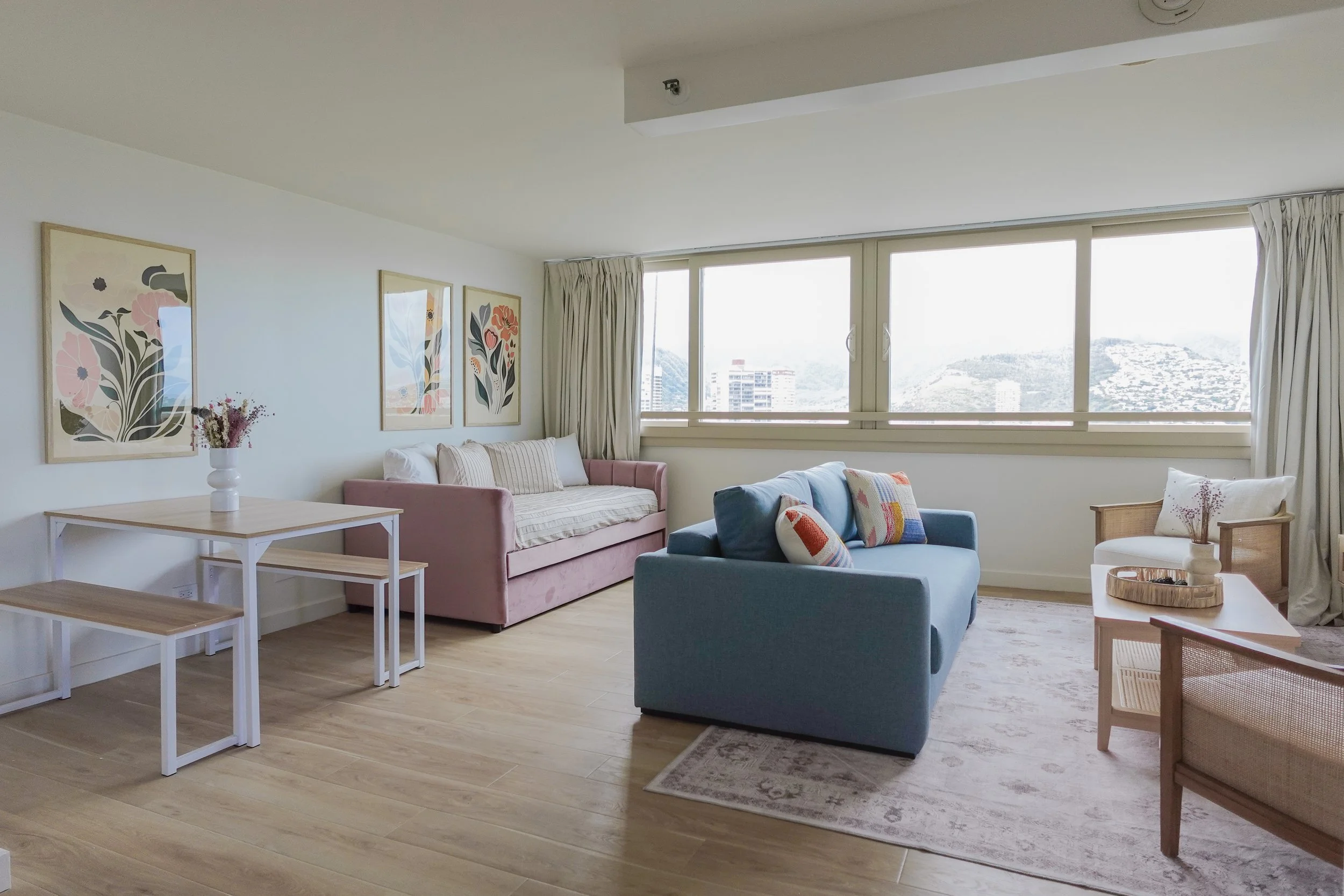 Modern living room with pink and blue sofas, wooden table with benches, floral artwork on walls, large windows with city view, and light wooden floors.