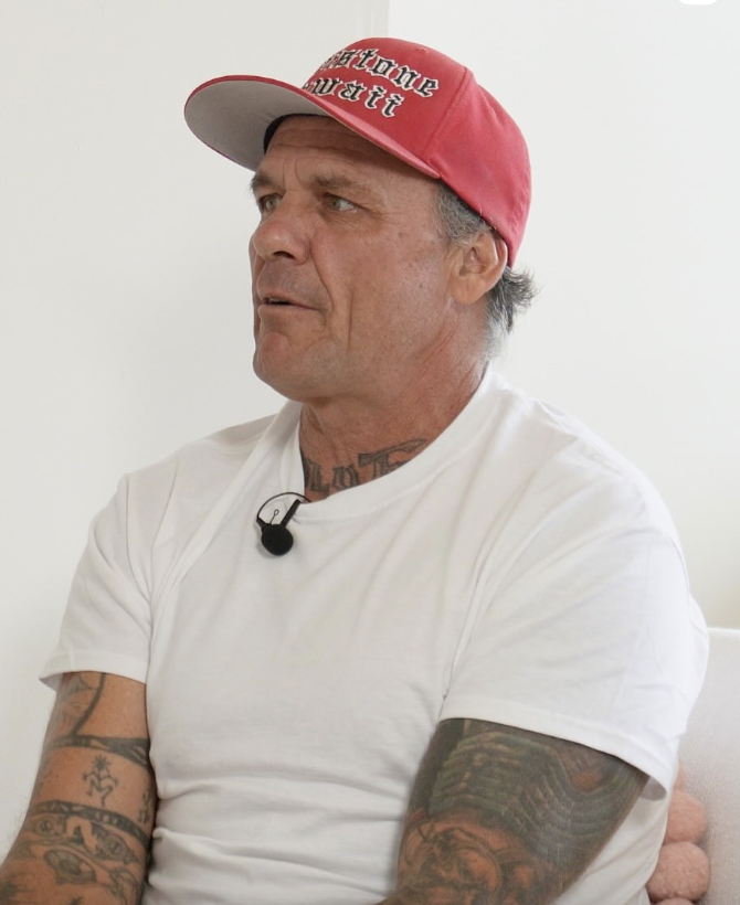 Man with tattoos wearing a red cap and white shirt, speaking or being interviewed, sitting indoors.