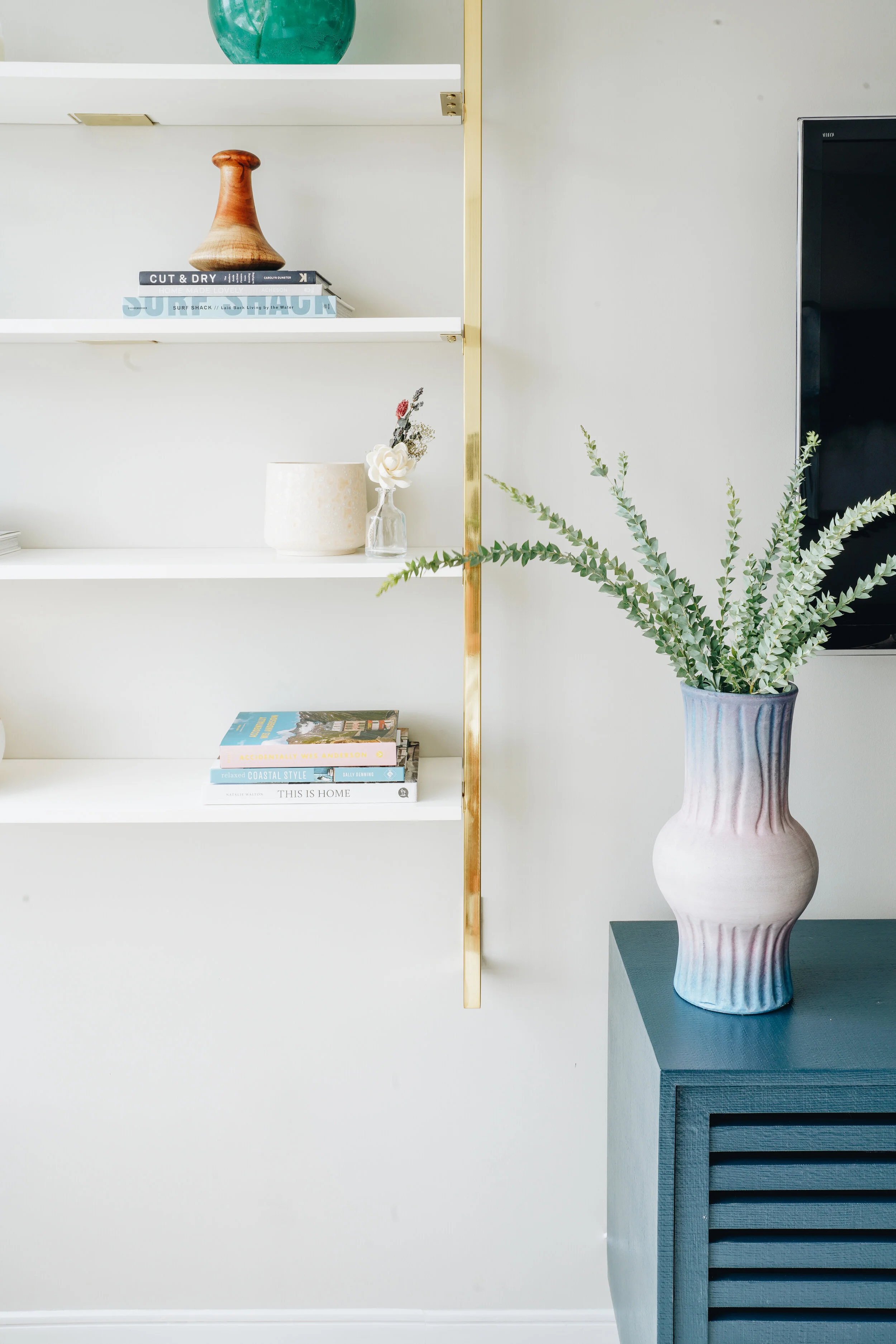 Living room with a decorative shelf, featuring books, a wooden vase, a green ceramic pot, and small decorative items. A large ceramic vase with greenery is on a blue console table, and a flat-screen TV is mounted on the wall.