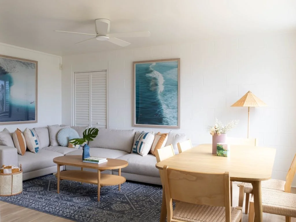 Modern coastal living room with a gray sectional sofa, wooden coffee table, blue patterned rug, and dining area. Decor includes ocean-themed wall art, a potted plant, and a woven basket. Soft natural lighting.