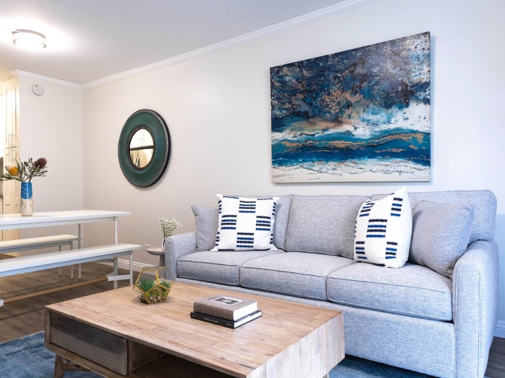 Modern living room with gray sofa, decorative pillows, abstract painting, round mirror, wooden coffee table, and a dining set.