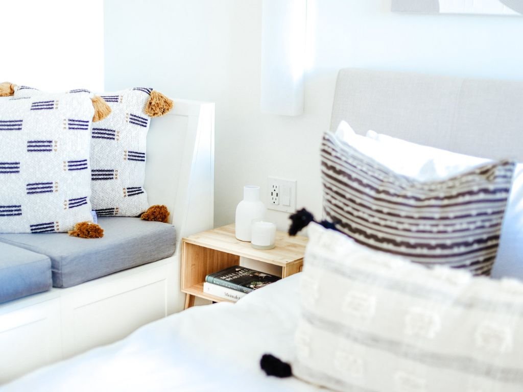 Cozy bedroom corner with patterned throw pillows, a small side table holding books and decor, and a soft bed with additional pillows.