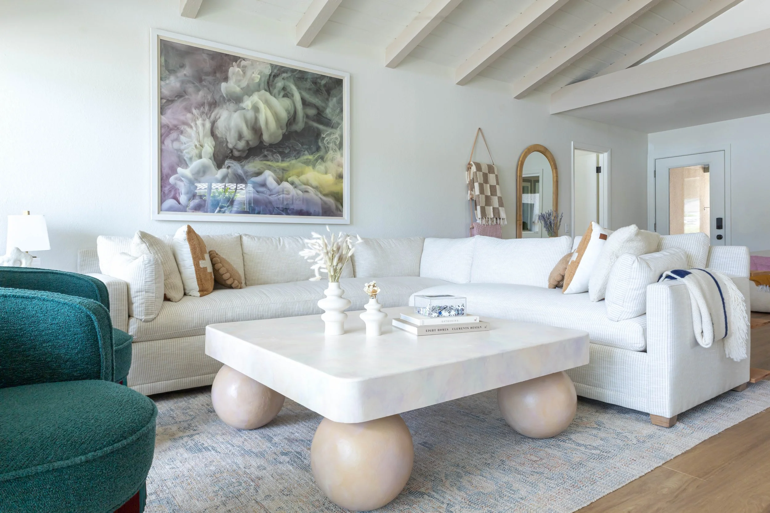 Modern living room with a white sectional sofa, a large abstract painting, a unique coffee table with ball-shaped legs, blue-green armchair, and decorative items on the table.