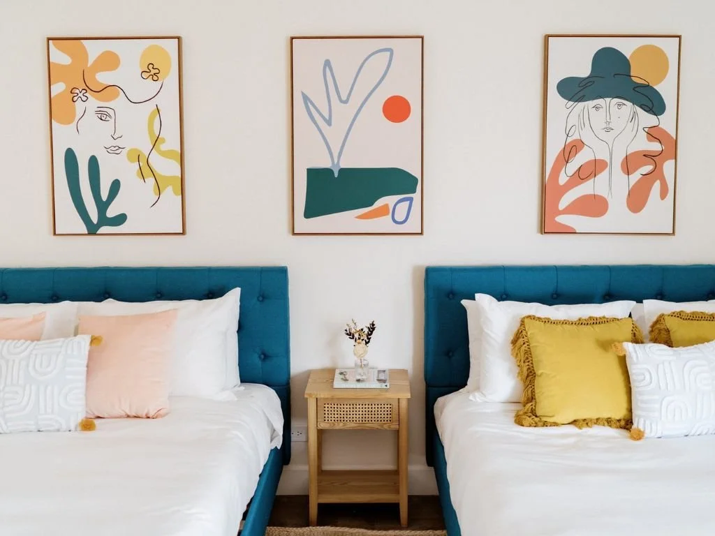 Bedroom interior with two teal upholstered beds, white bedding, yellow and peach pillows, a central wooden nightstand, and three abstract art pieces on the wall above the beds featuring colorful geometric shapes and line drawings.