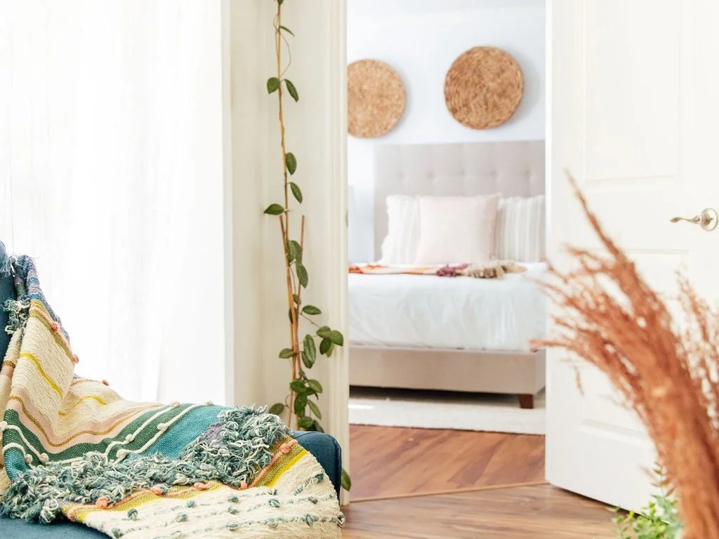 Decorative interior with a colorful throw blanket on a chair, a plant, woven wall hangings, and a doorway leading to a bedroom with light bedding.