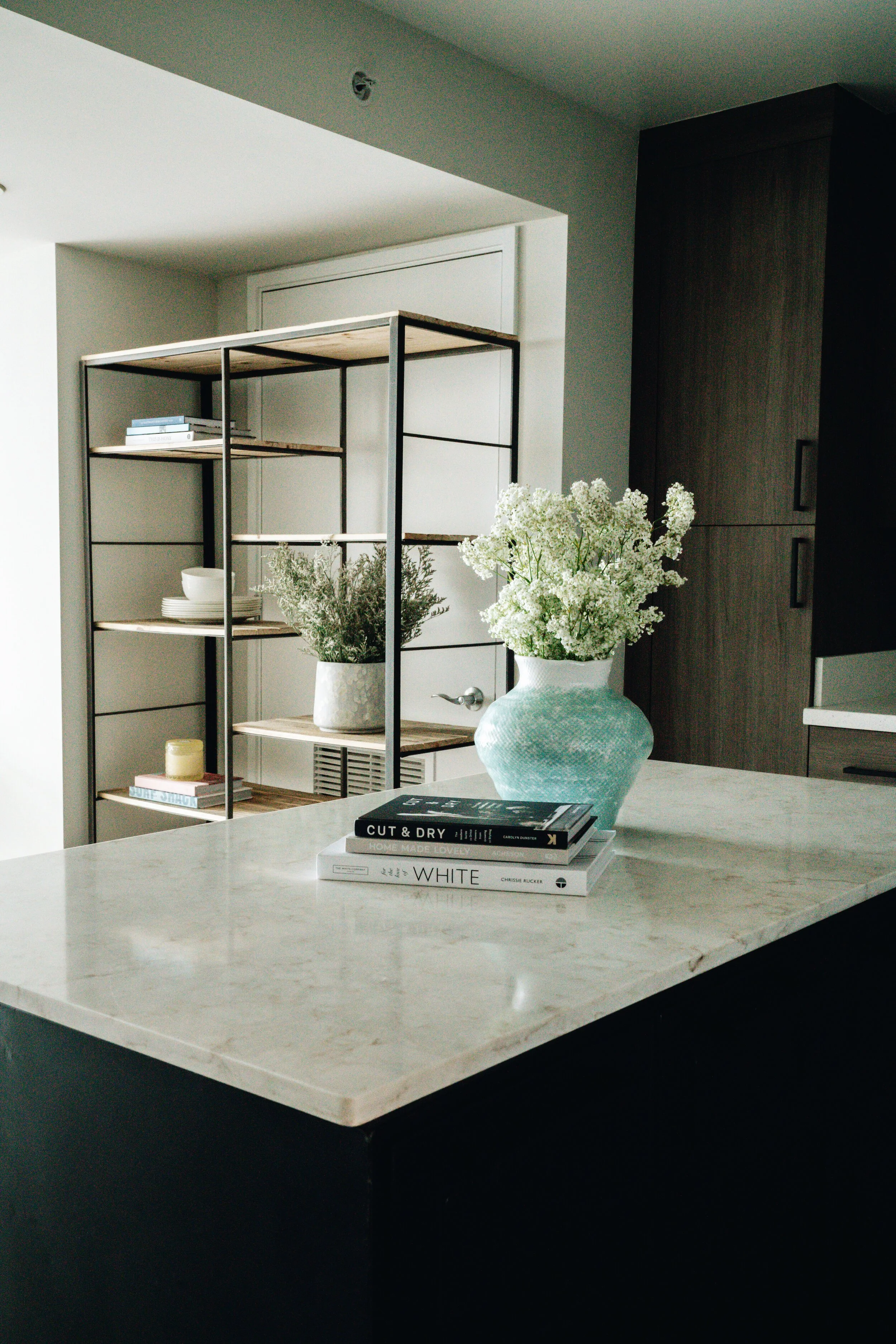 Modern kitchen interior with marble countertop, books, blue vase with white flowers, metal shelving unit, potted plant, and dishware.