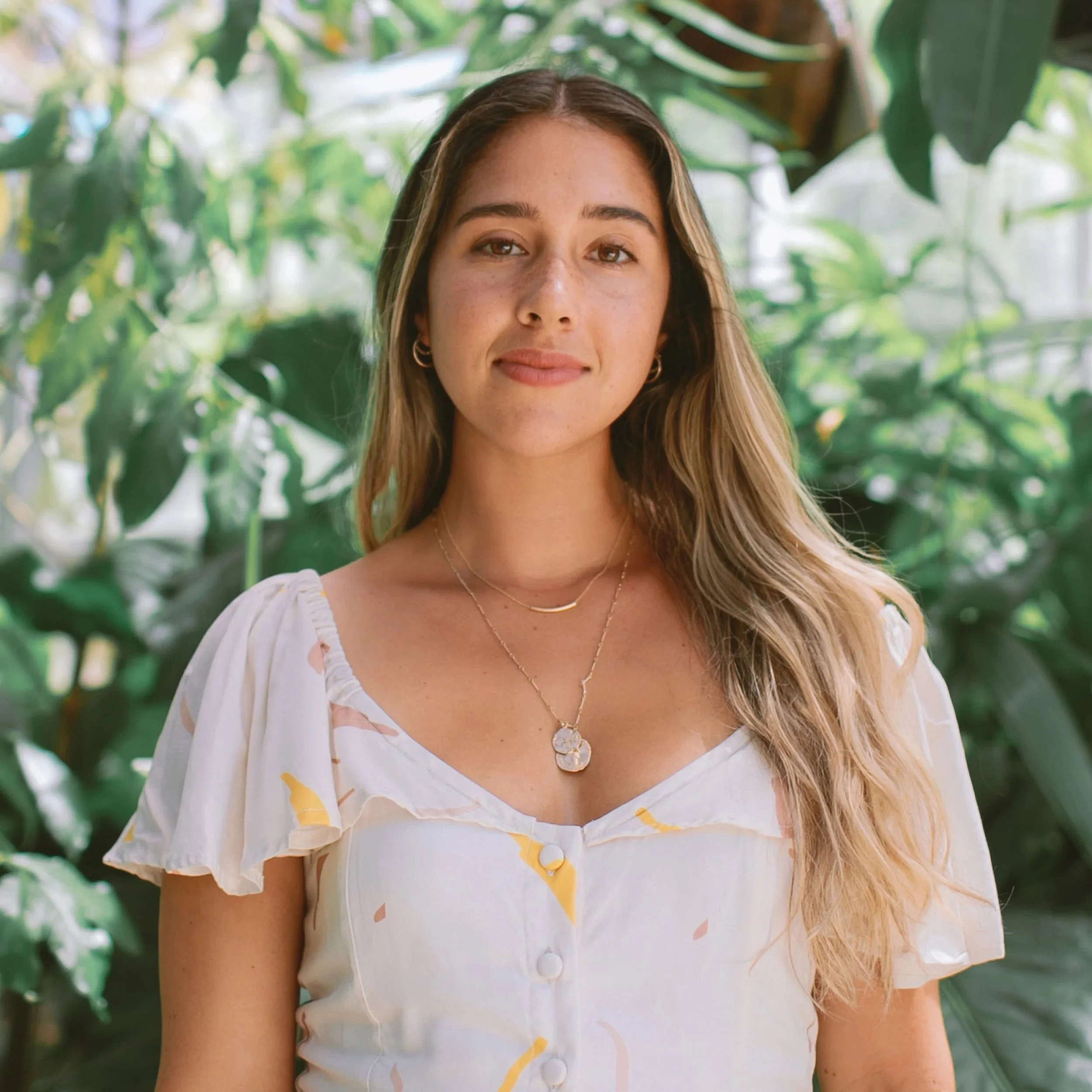 Woman standing in front of lush green foliage, wearing a light-colored dress with floral patterns and gold necklaces.