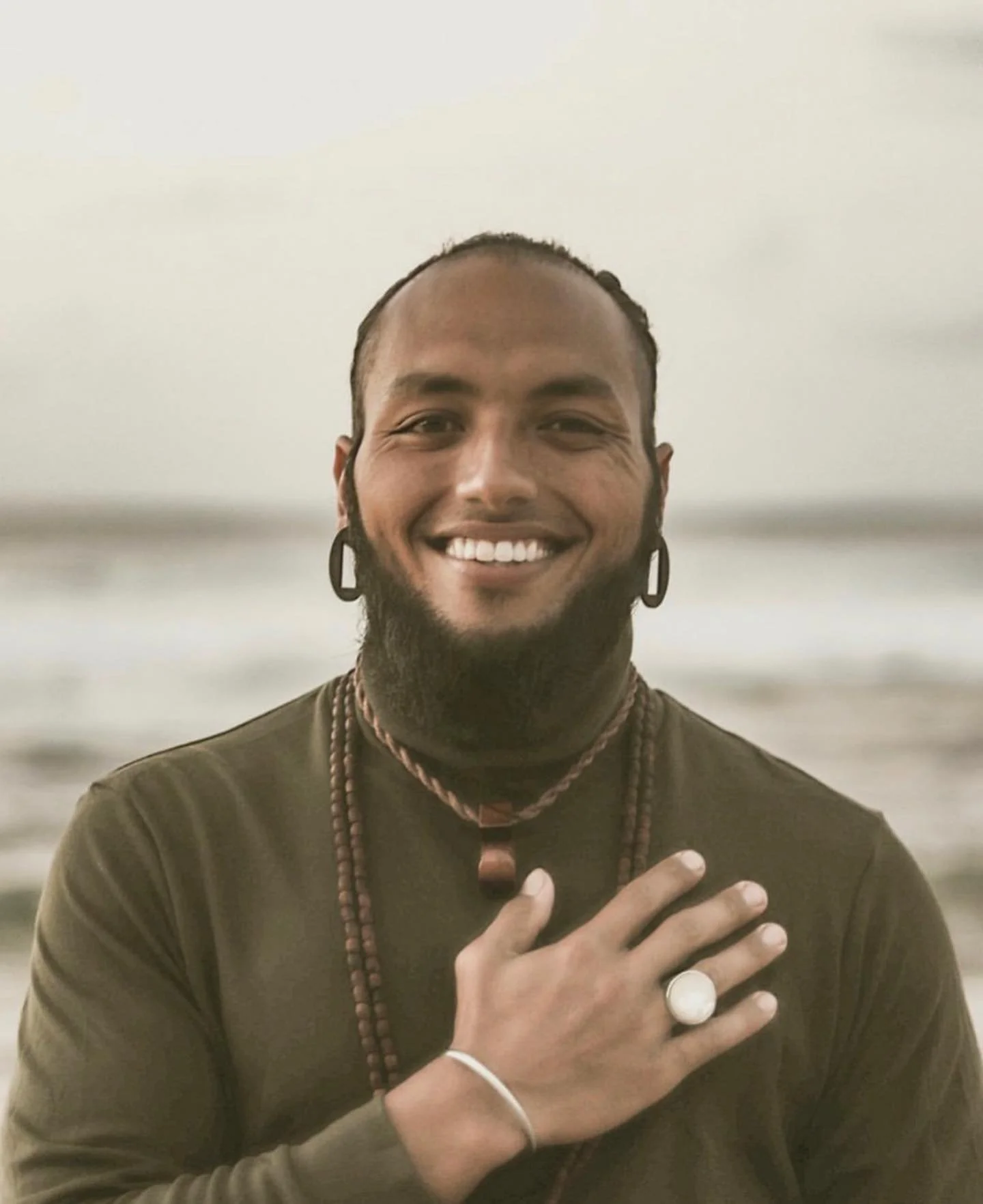 Smiling person with beard and earrings facing the camera, wearing a green shirt and necklaces, outdoors.