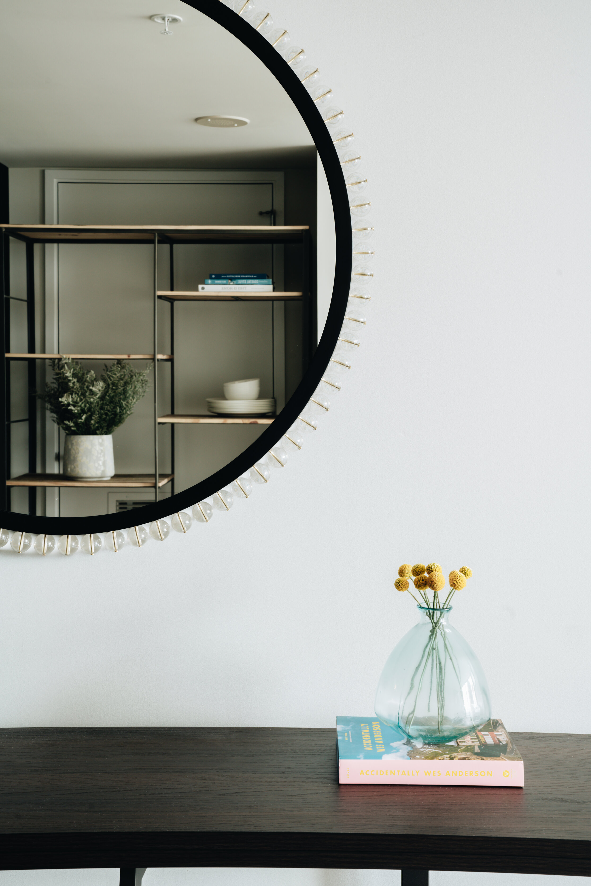 Round mirror reflecting bookshelves; table with a vase of yellow flowers on top of a book titled "Accidentally Wes Anderson."