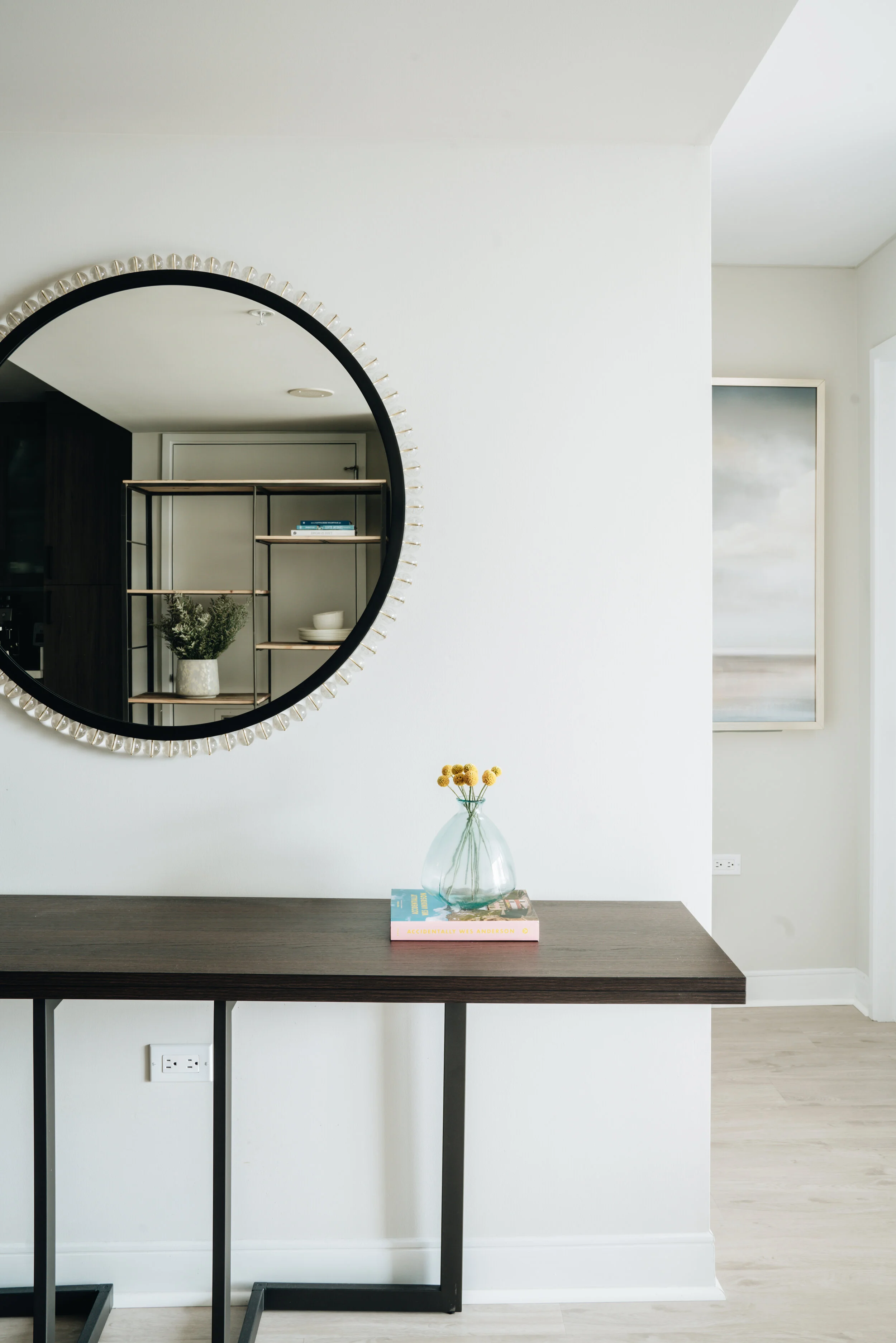 Modern minimalist entryway with round mirror, dark console table holding a small vase with yellow flowers, and a stack of books.