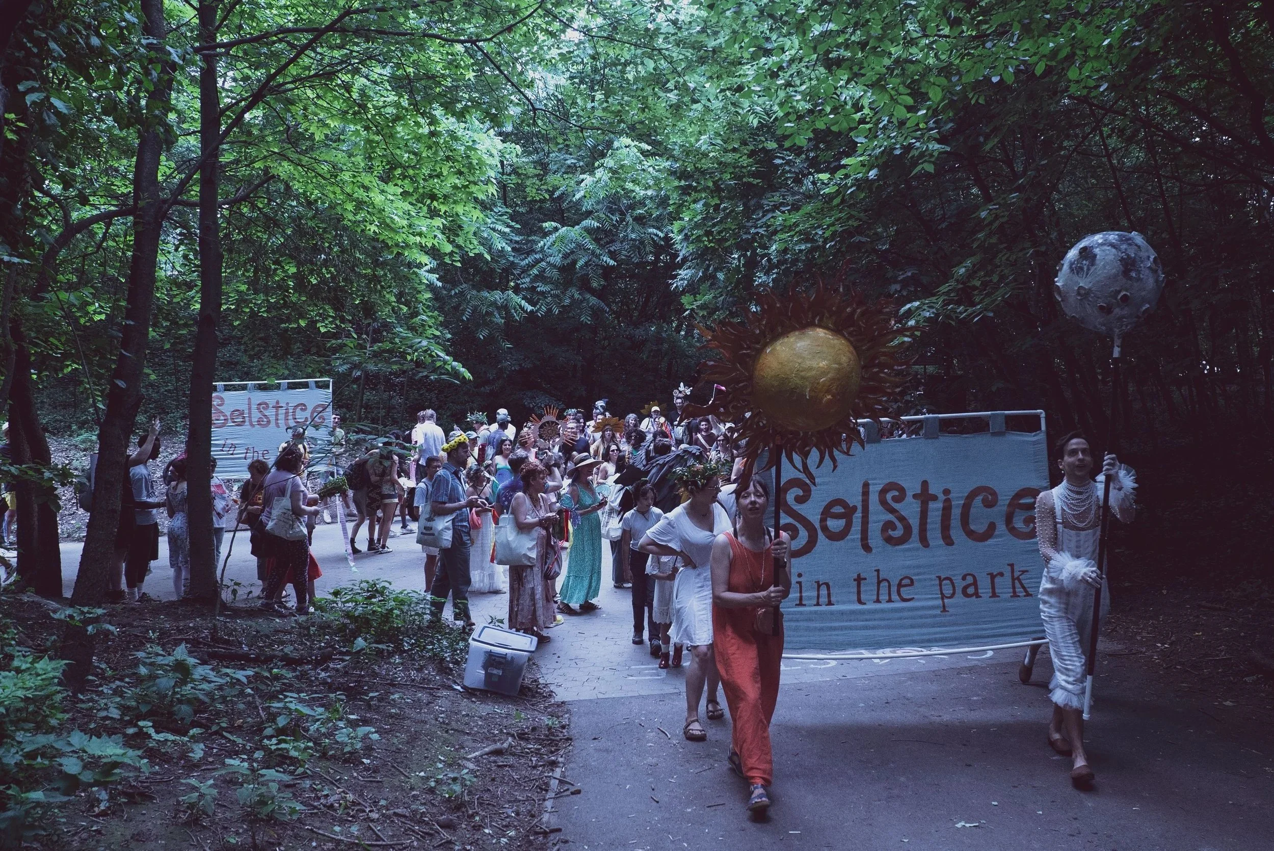 Two people carry large circular puppets through a path in the forest. They are followed by a group of people walking.