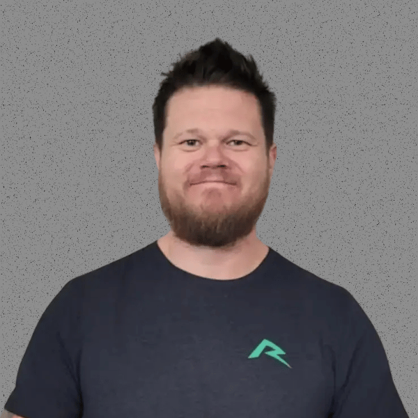 Man with a beard and short dark hair smiling, wearing a navy blue t-shirt with a teal logo, standing against a plain gray background.