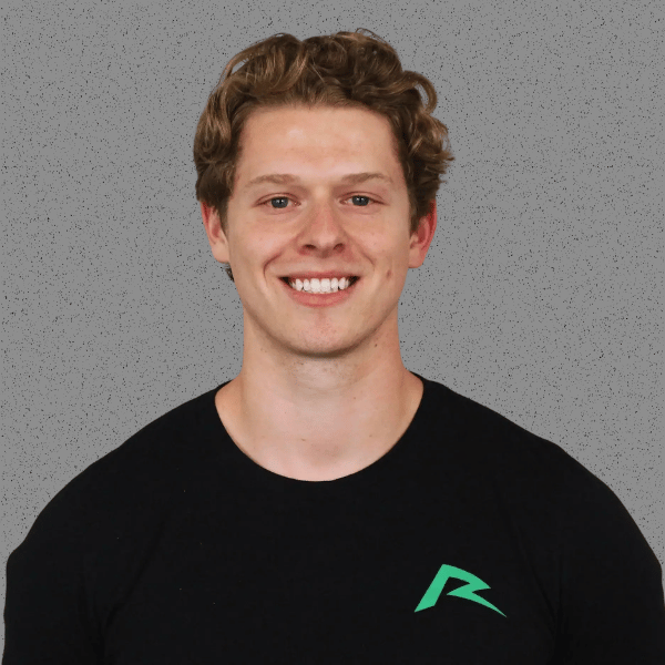 A young man with curly brown hair and a big smile, wearing a black T-shirt with a green logo on the chest, standing in front of a gray background.