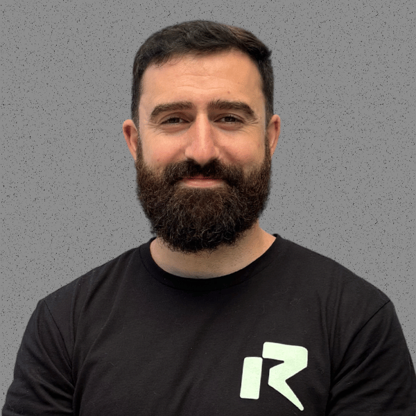 A man with a beard and short dark hair standing against a gray background, wearing a black t-shirt with a green logo.