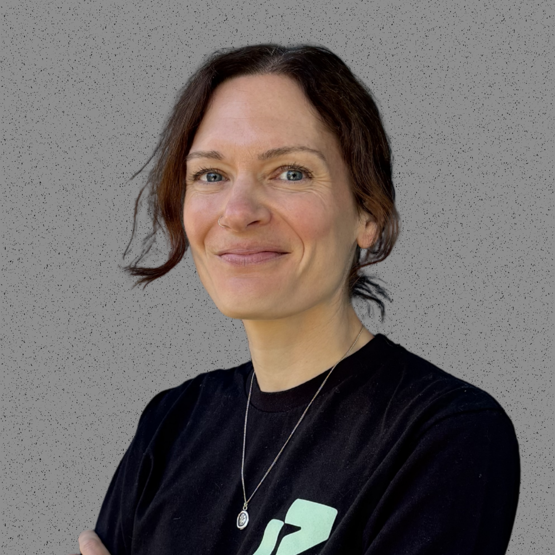 Close-up of a smiling woman with short brown hair, blue eyes, wearing a black shirt with a logo, a necklace with a pendant, and a nose ring, standing against a textured grey background.