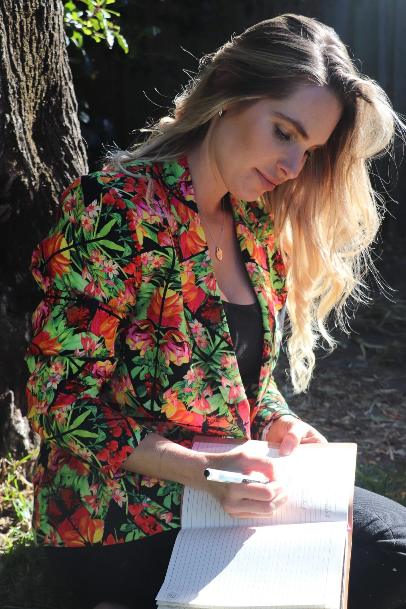 A woman with long, wavy blonde hair, wearing a colorful floral blazer, is sitting outside near a tree, writing in a notebook with a black pen.