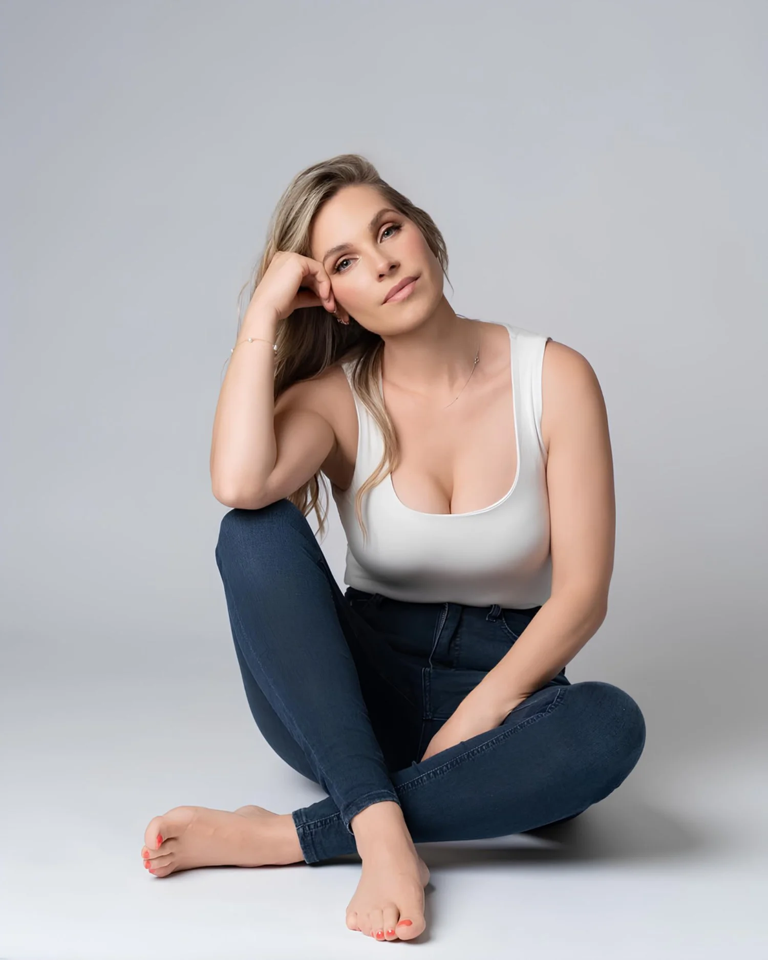 A woman with long wavy blonde hair sitting on the floor, wearing a white tank top and dark blue jeans, with one hand resting on her head and a neutral expression.