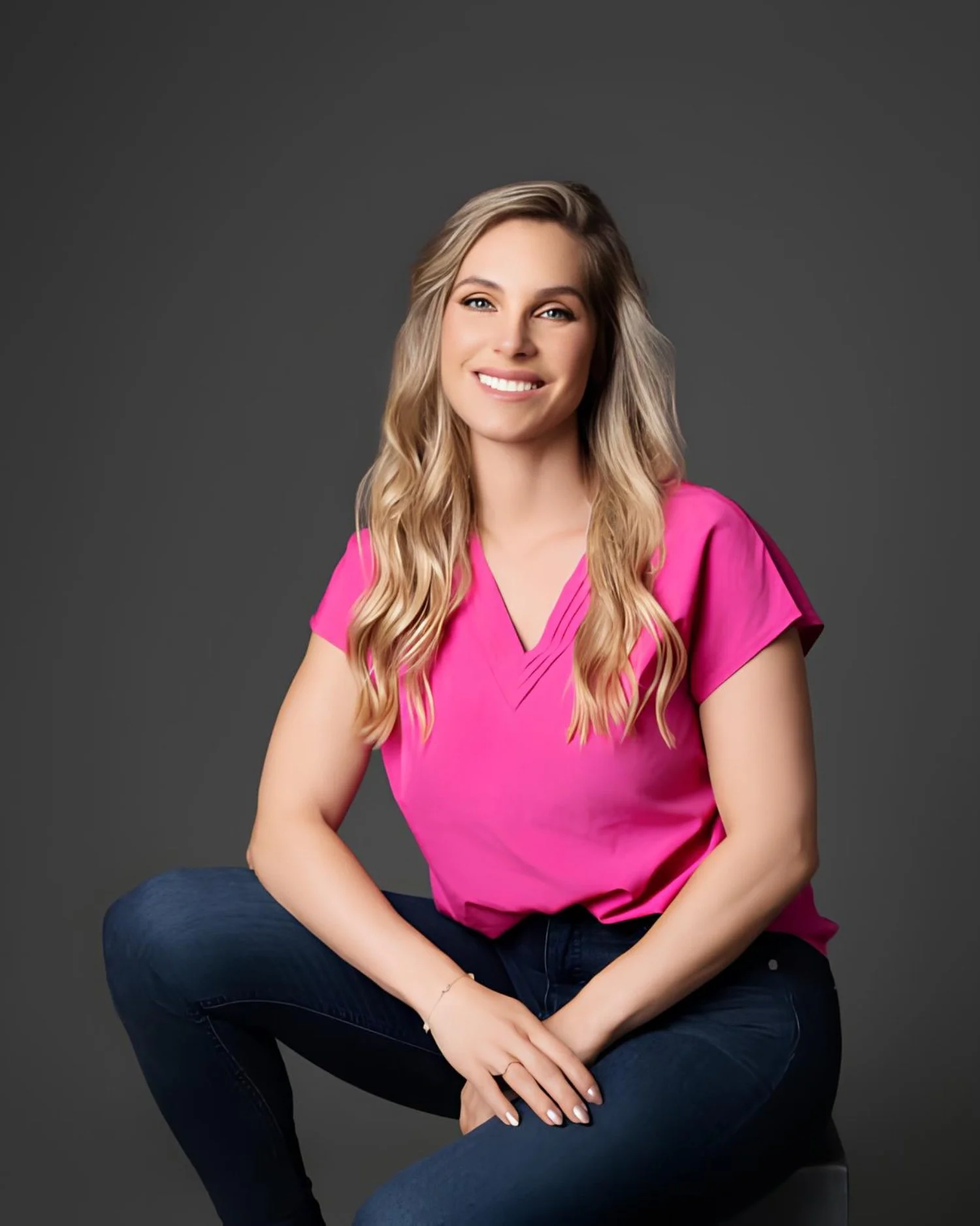 A smiling woman with long blonde wavy hair wearing a pink blouse and dark jeans, sitting against a dark gray background.