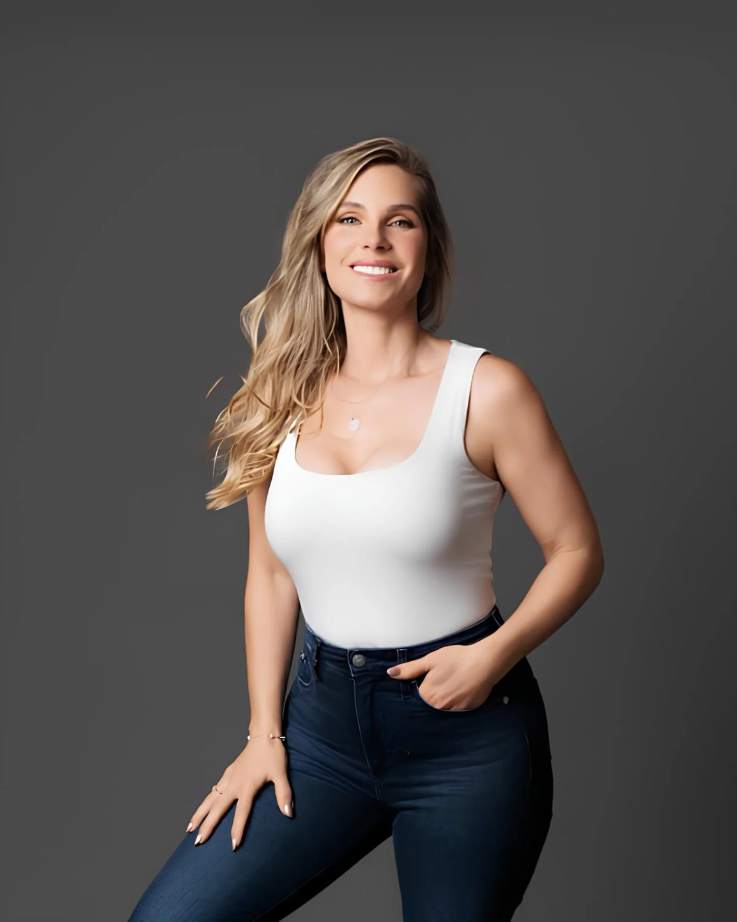 A smiling woman with long blonde hair, wearing a white tank top and dark blue jeans, standing against a gray background.